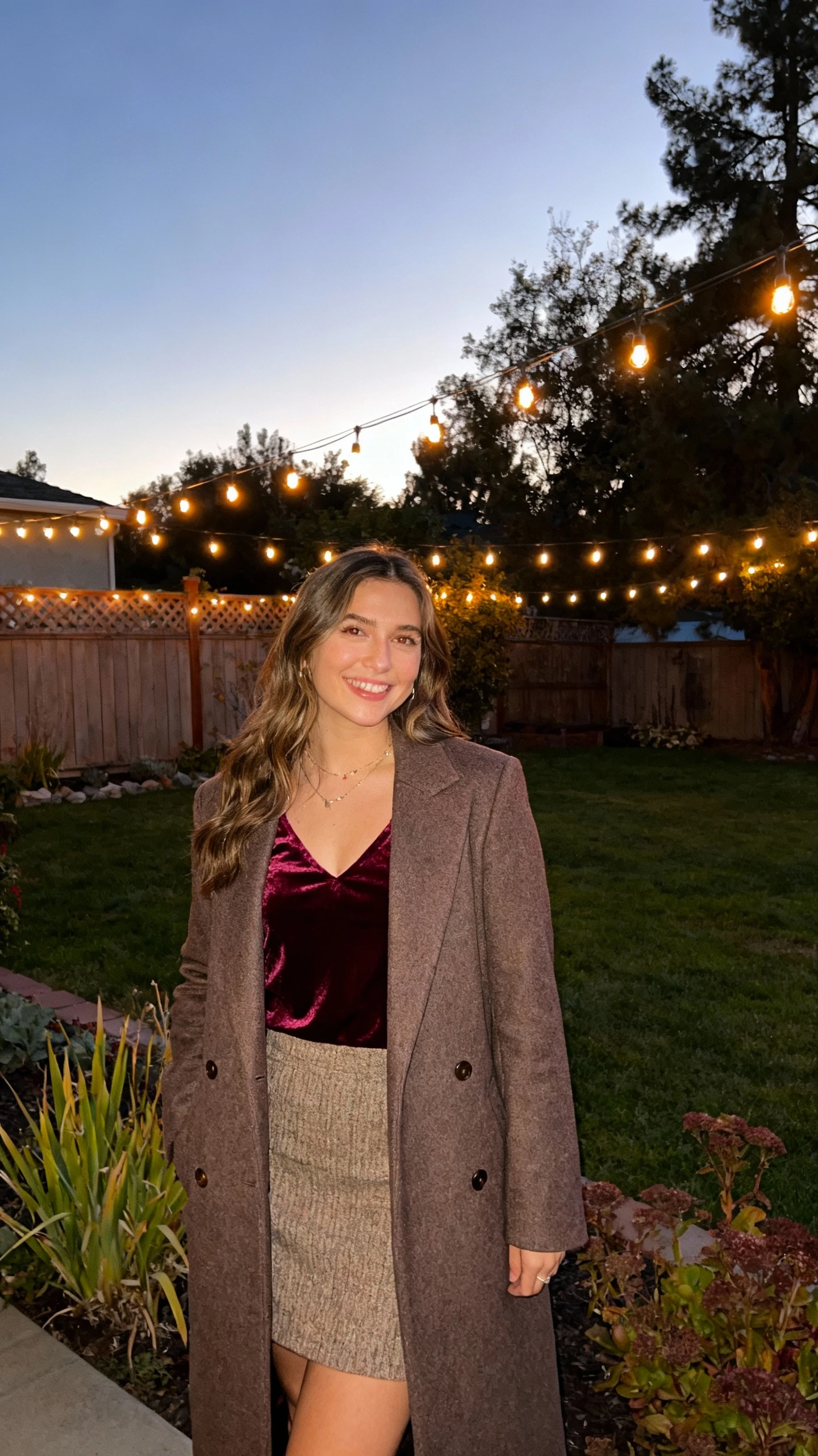 A woman dressed for evening in a velvet top with a wool skirt and tailored wool coat, standing near twinkling garden lights at dusk in a backyard, casual iPhone photo style, natural daylight, outdoor setting.