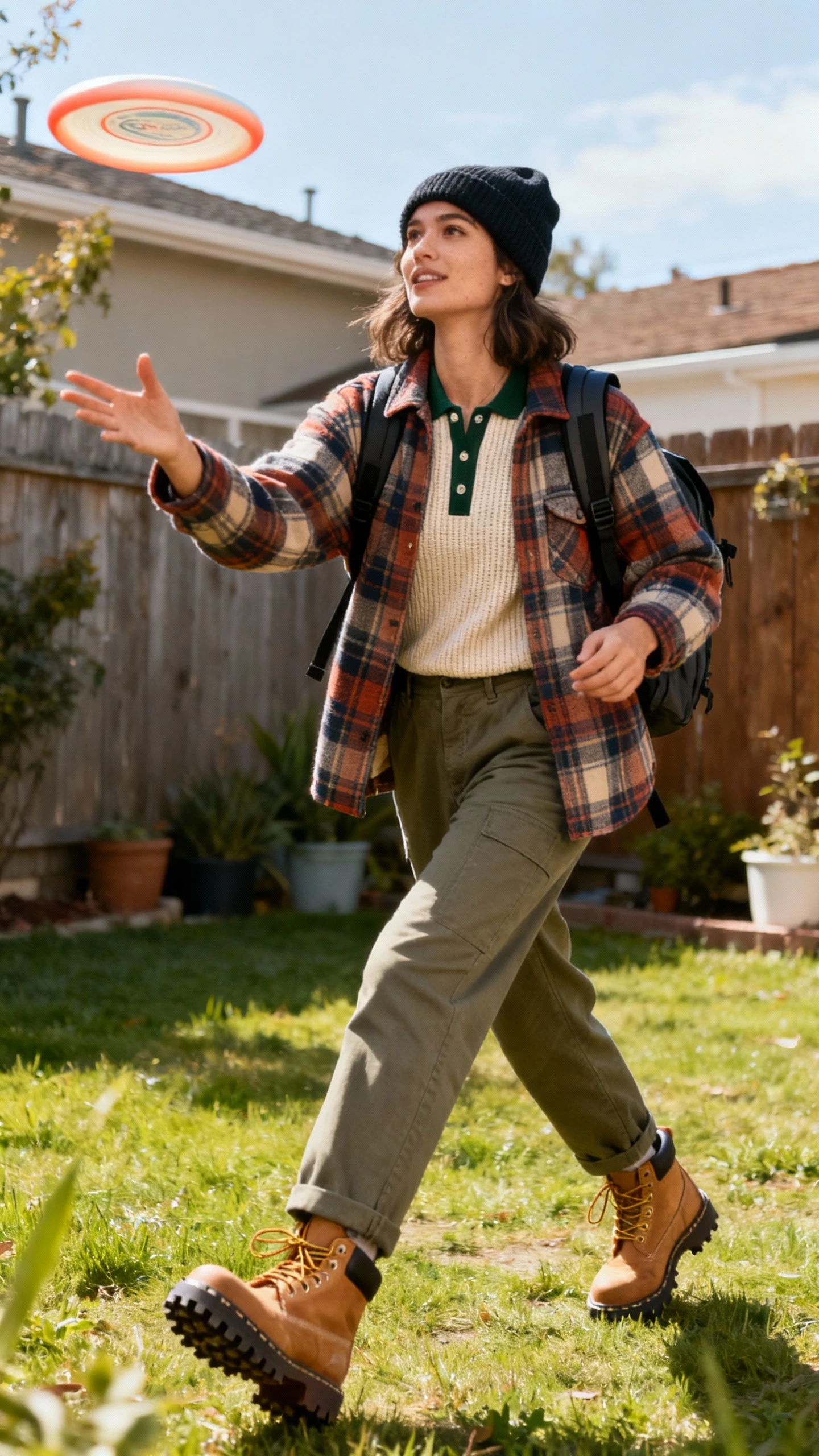 A woman wearing a plaid shacket layered over a waffle-knit henley with straight-leg cargos and lug-sole boots, beanie and backpack, tossing a frisbee in a backyard, casual iPhone photo style, natural daylight, outdoor setting, easygoing movement.