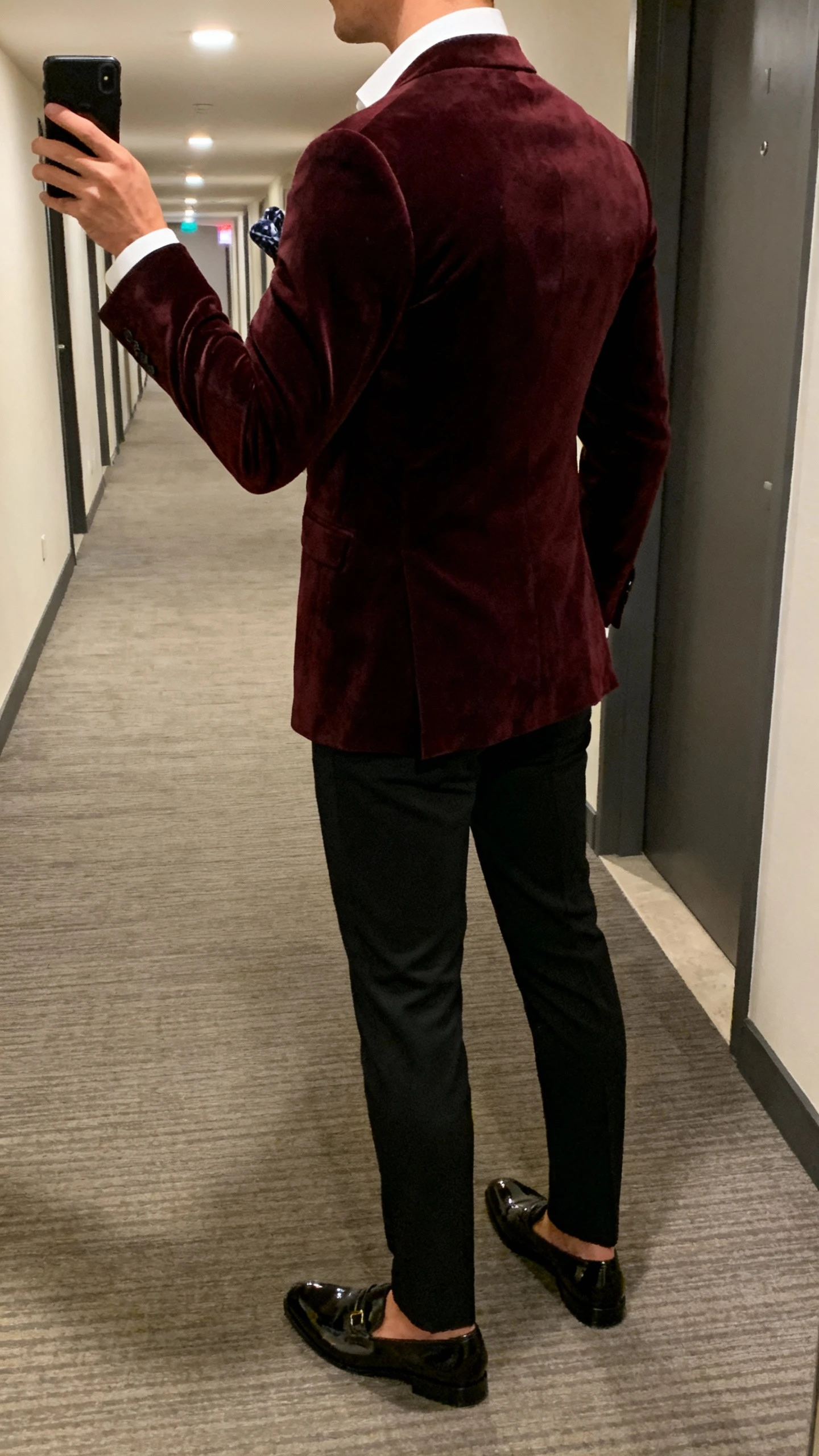iPhone back-view hallway selfie of a man in a deep burgundy velvet blazer over a crisp white shirt, black dress trousers, patent loafers, and a pocket square, face not visible, hotel corridor lighting, casual iPhone quality.