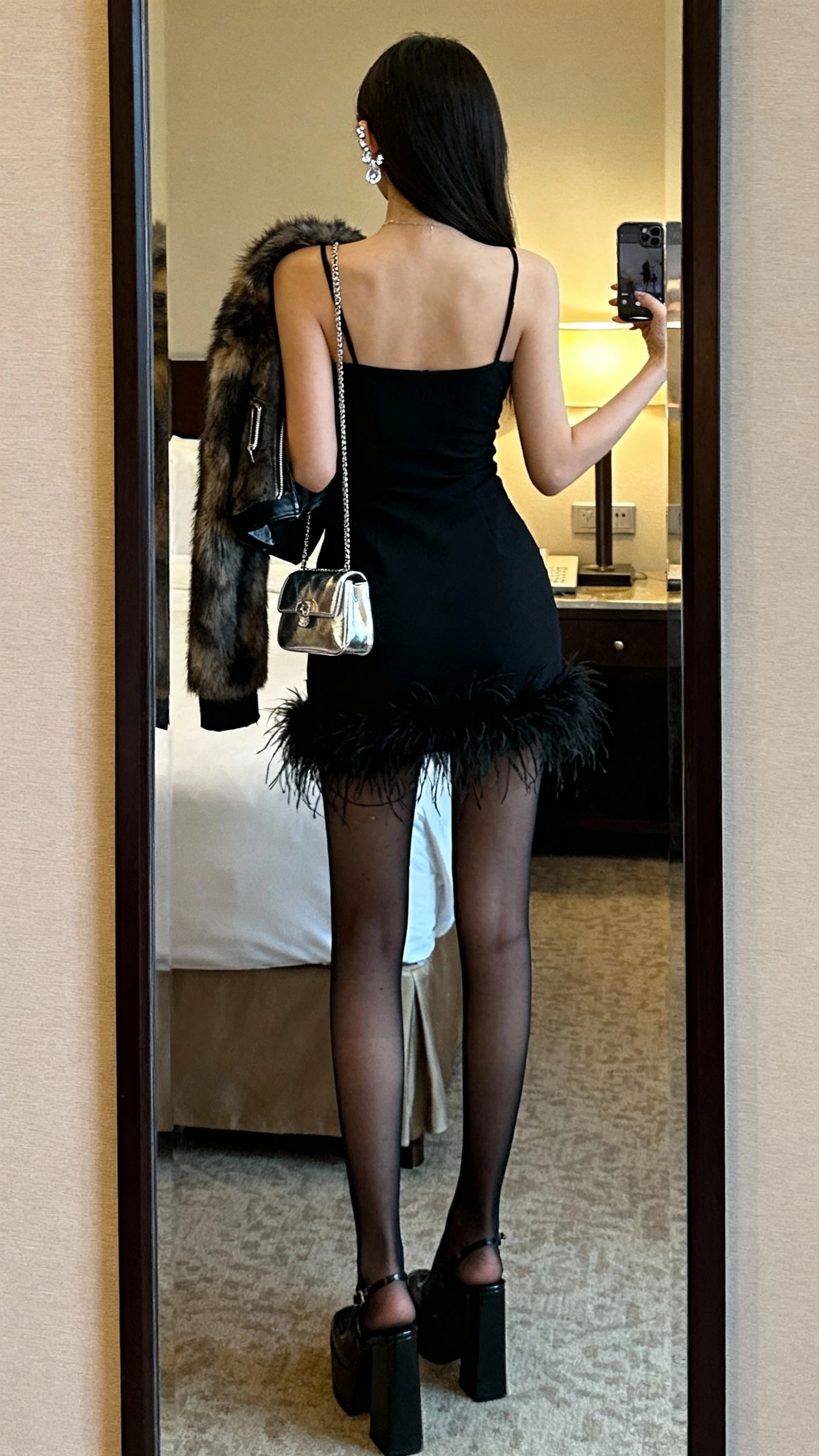 iPhone back-view mirror selfie of a woman in a black feather-trim mini dress, sheer black tights, platform heels, metallic micro bag, crystal ear climbers, and a cropped faux-fur jacket slung over one shoulder; face not visible, hotel room mirror, ambient lamp light, natural iPhone photo quality.
