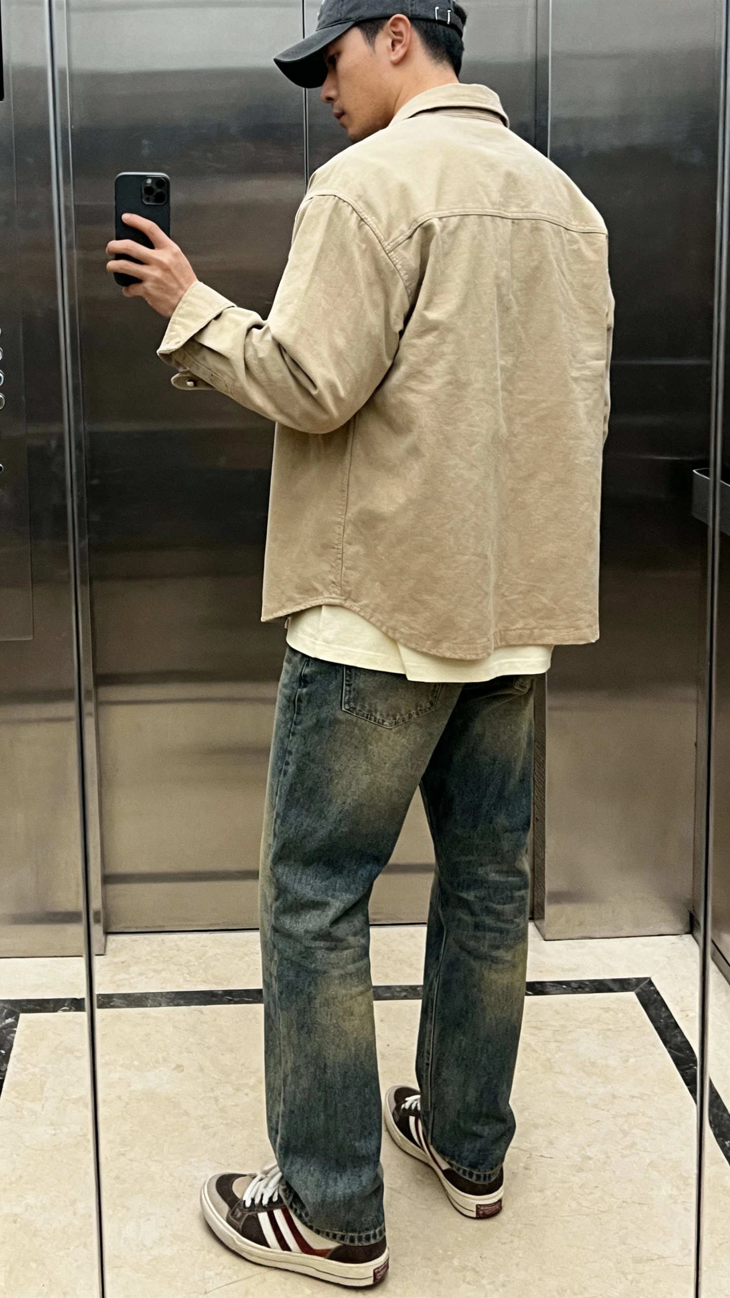 iPhone back-view selfie of a man in a beige cotton-twill overshirt over a cream tee, straight-leg mid-wash denim, retro runners, and a charcoal cap; elevator mirror, indoor lighting, face not visible, casual iPhone photo.