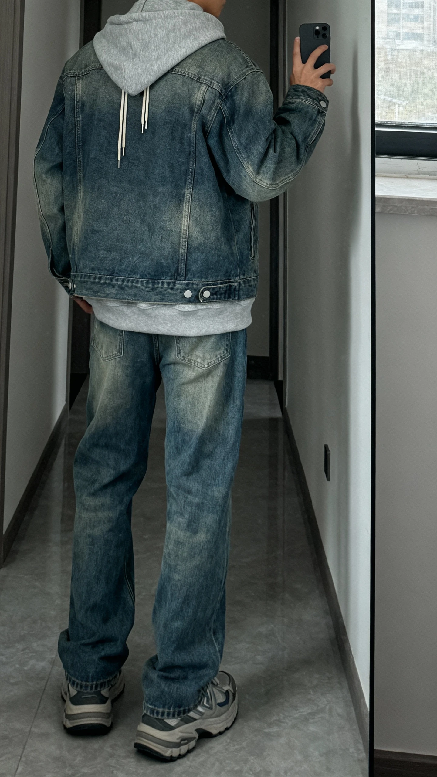 iPhone back-view selfie of a man wearing a medium wash denim jacket layered over a gray hoodie with drawstrings, relaxed blue jeans, and chunky sneakers, face not visible, hallway mirror, overcast daylight from window, casual iPhone photo.