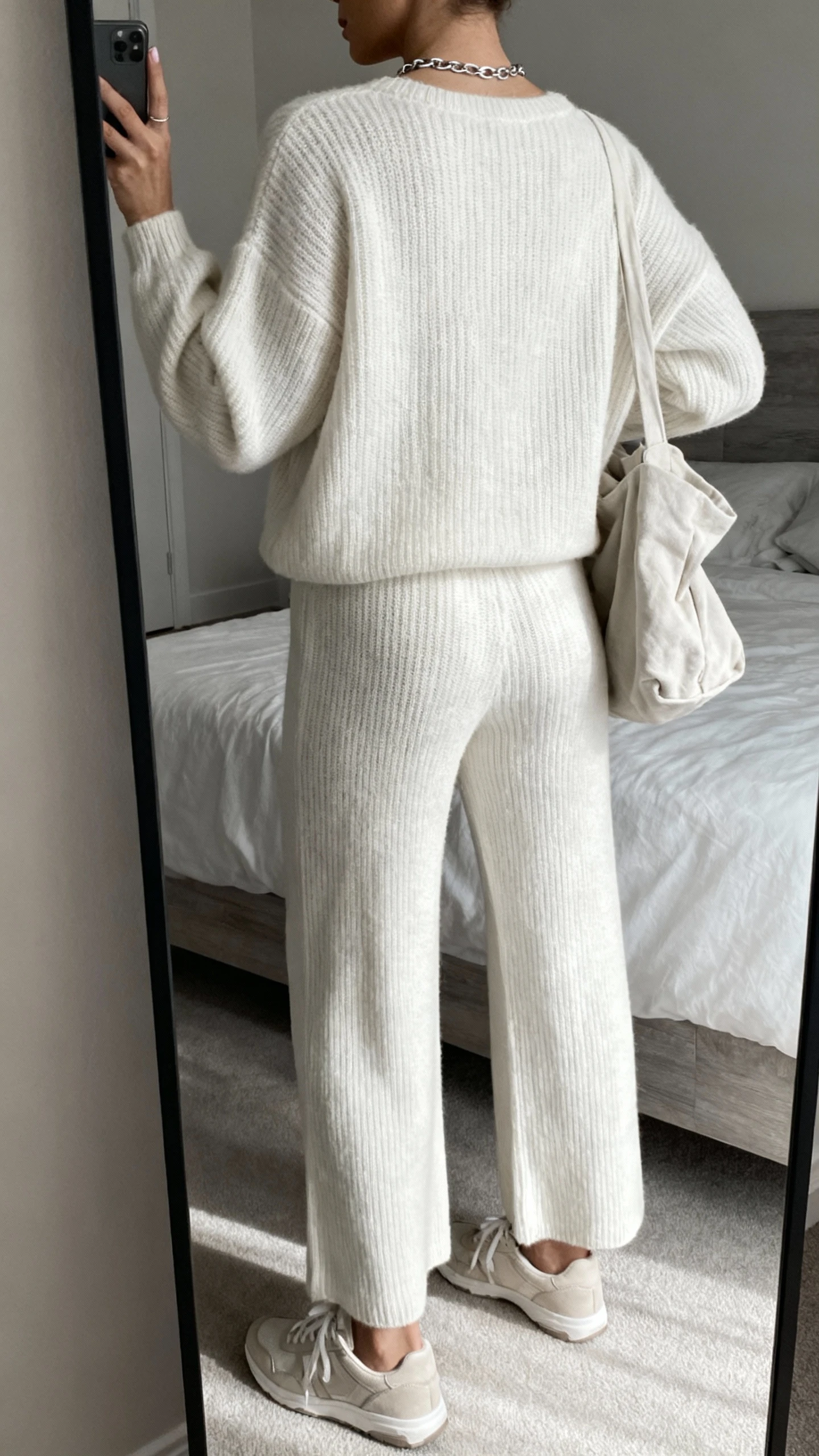 iPhone back-view selfie of a woman in a monochrome knit set (matching sweater and wide-leg knit pants), tonal sneakers, delicate chain necklace, and a slouchy tote, face not visible, bedroom full-length mirror, soft morning light, natural iPhone photo quality.