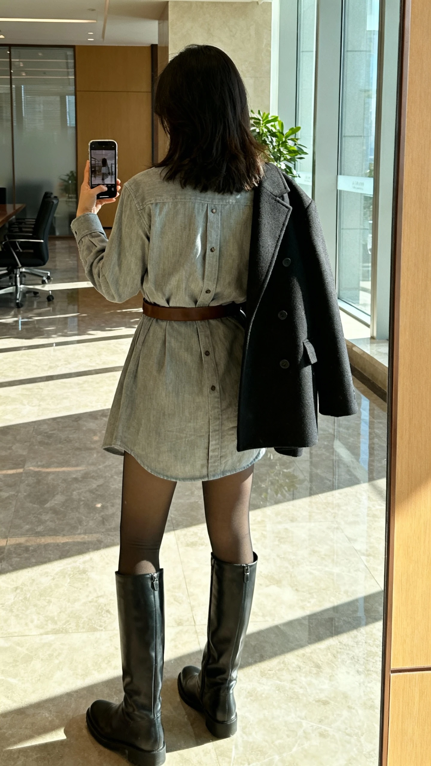 iPhone back-view selfie of a woman in a winterized shirt dress belted at the waist with tall knee boots, opaque tights, and a structured coat draped over shoulders, face not visible, office lobby mirror, bright morning light, natural iPhone photo quality.