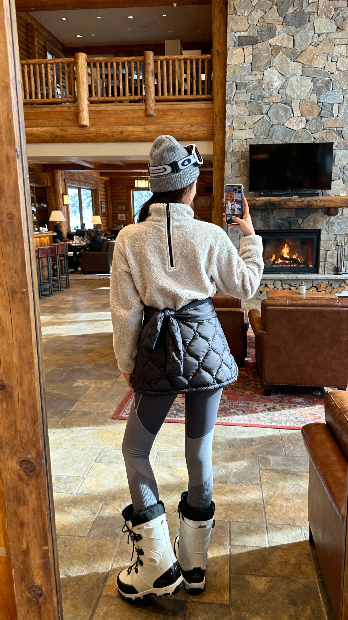 iPhone back-view selfie of a woman in an après-ski off-duty outfit: fleece half-zip, thermal leggings, chunky snow boots, quilted vest tied at waist, beanie, ski-style sunglasses on head, face not visible, lodge lobby near fireplace, warm indoor lighting, natural iPhone quality.