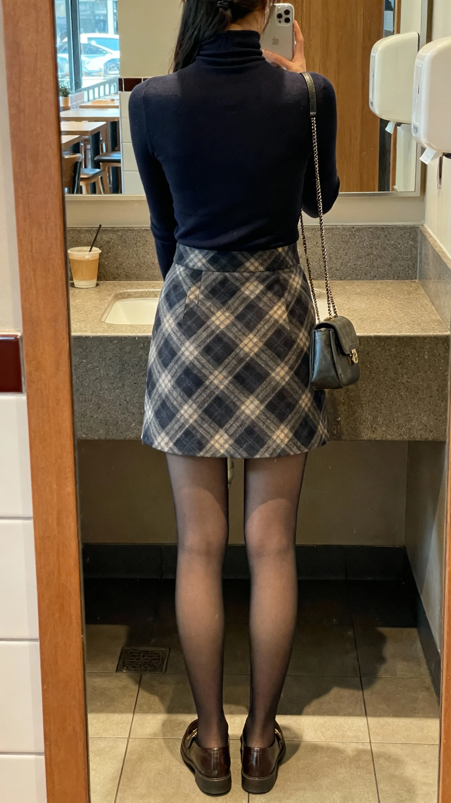iPhone back-view selfie of a woman wearing a plaid mini skirt, fitted turtleneck, opaque tights, loafers, small shoulder bag, face not visible, cafe restroom mirror, soft indoor lighting, casual iPhone aesthetic.