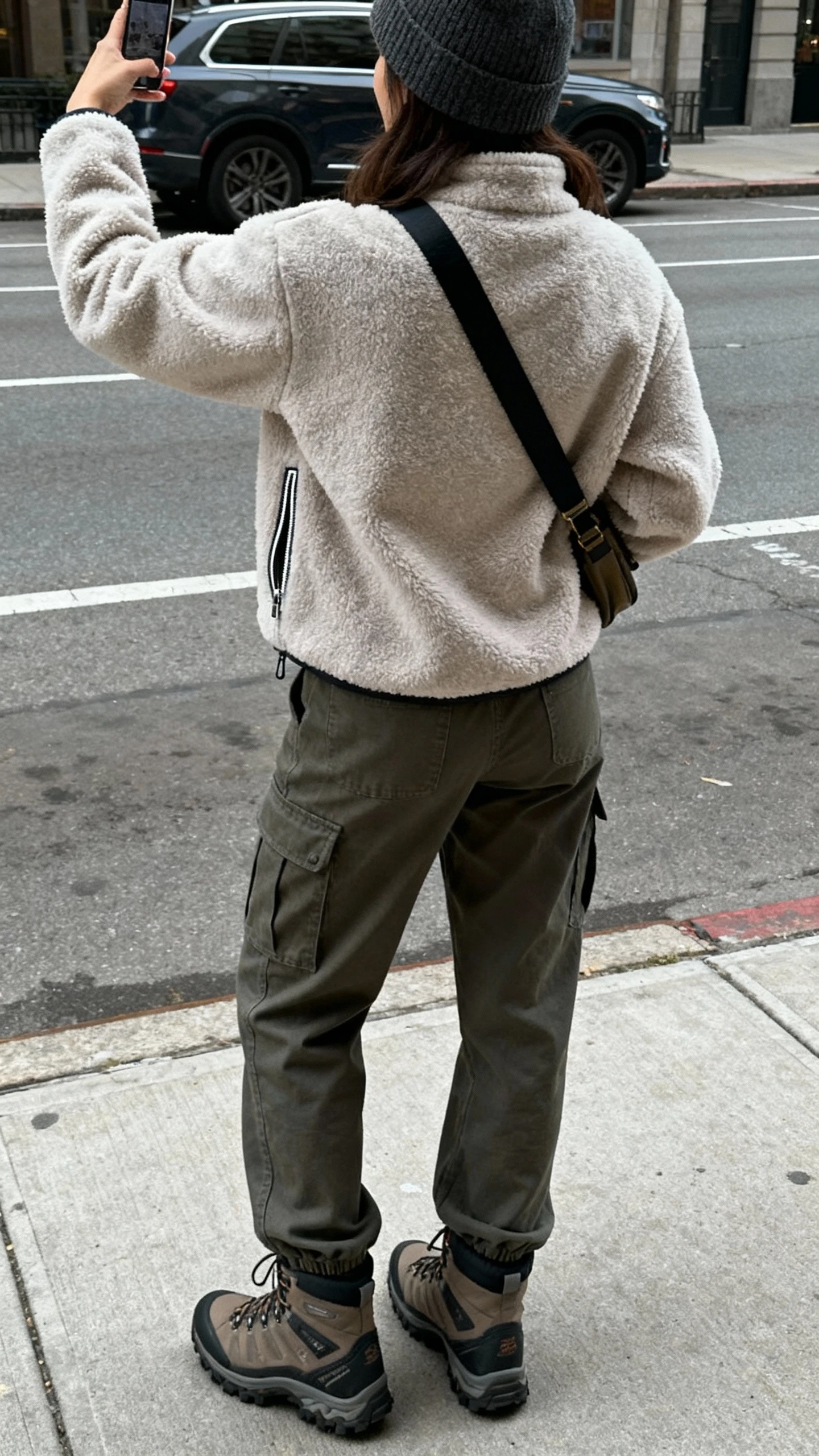 iPhone back-view selfie of a woman wearing cargo pants, a fleece zip-up, city hiker boots, and a beanie, crossbody bag on, face not visible, urban sidewalk with overcast daylight, casual iPhone photo.