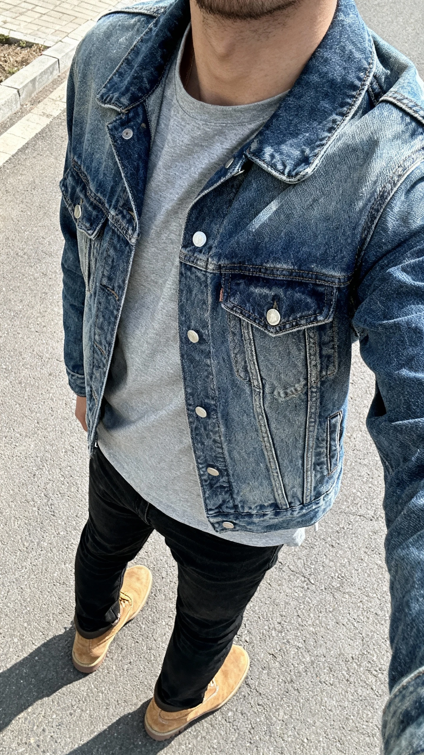 iPhone back-view street selfie of a man wearing a mid-wash denim jacket over a gray tee with black jeans and desert boots, face not visible, over-the-shoulder angle in daylight, casual iPhone photo.
