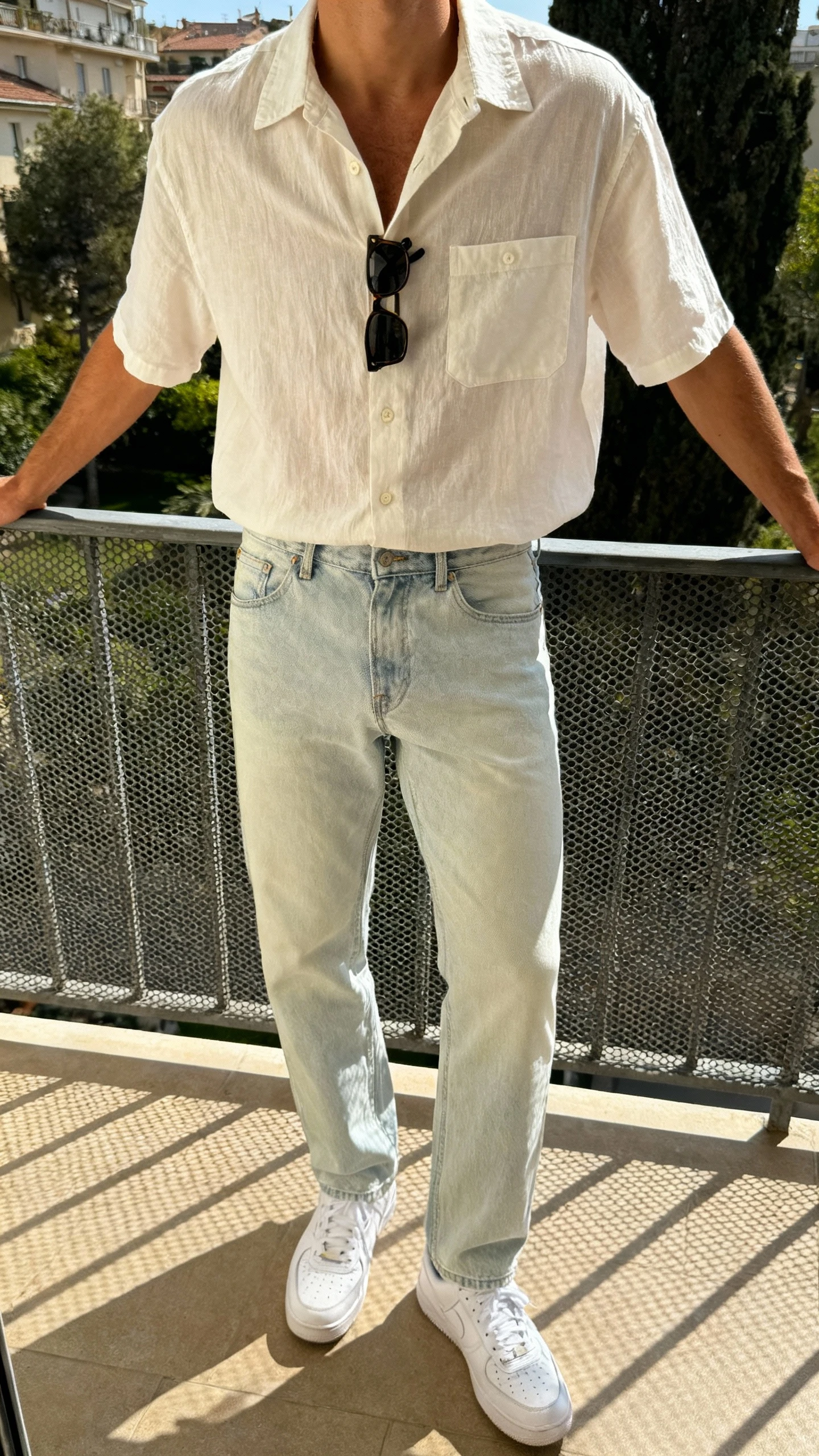 iPhone balcony selfie cropped at neck showing a man in a breezy short-sleeve button-up (camp-collar) tucked into lightweight light-wash jeans with white low-top sneakers and sunglasses clipped to the placket, face not visible, sunny natural light, casual iPhone photo.