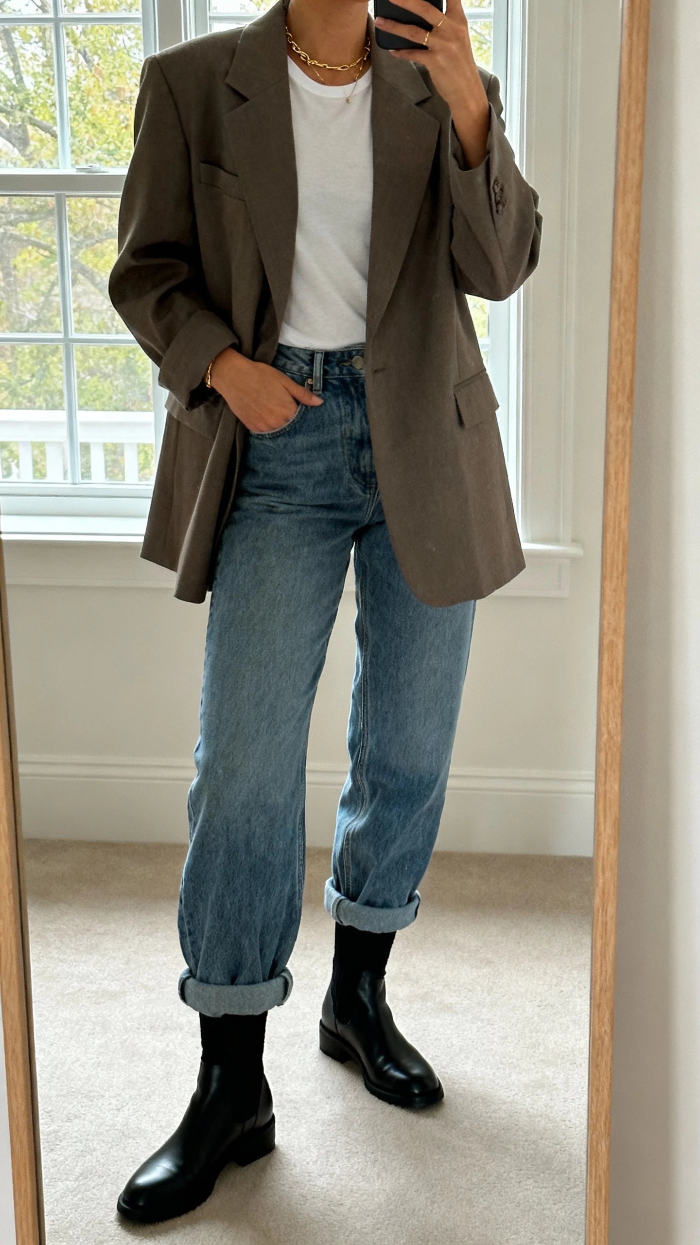 iPhone bedroom mirror selfie of a woman in an oversized blazer, barrel-leg jeans, sleek black sock boots, fitted tee, and delicate gold jewelry; face not visible, full-length mirror, soft afternoon window light, natural iPhone photo quality.
