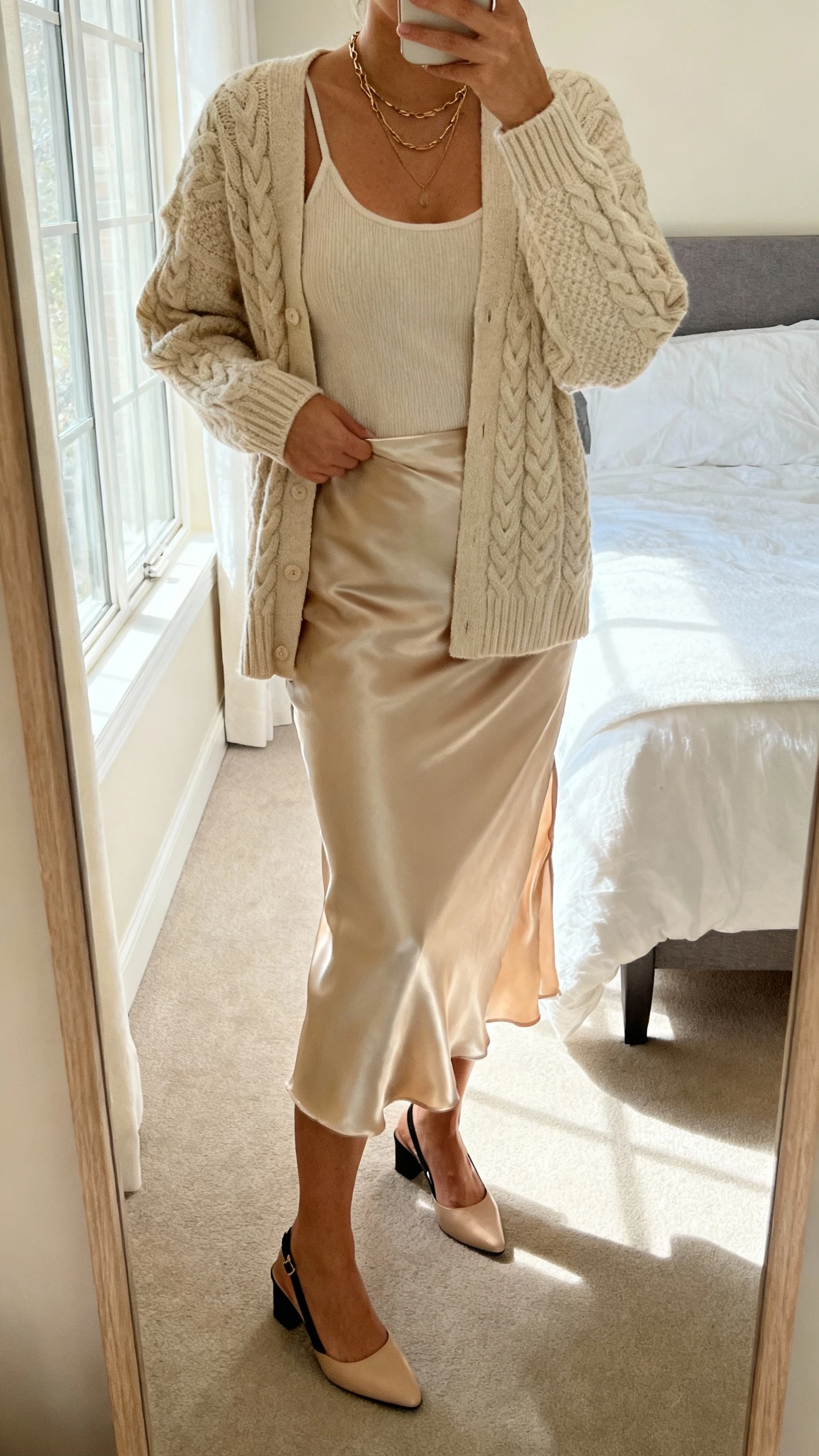 iPhone bedroom mirror selfie of a woman wearing a cable-knit cardigan set (cardigan + knit tank) with a satin slip skirt, slingback flats, layered necklace, face not visible, morning window light, natural iPhone look.