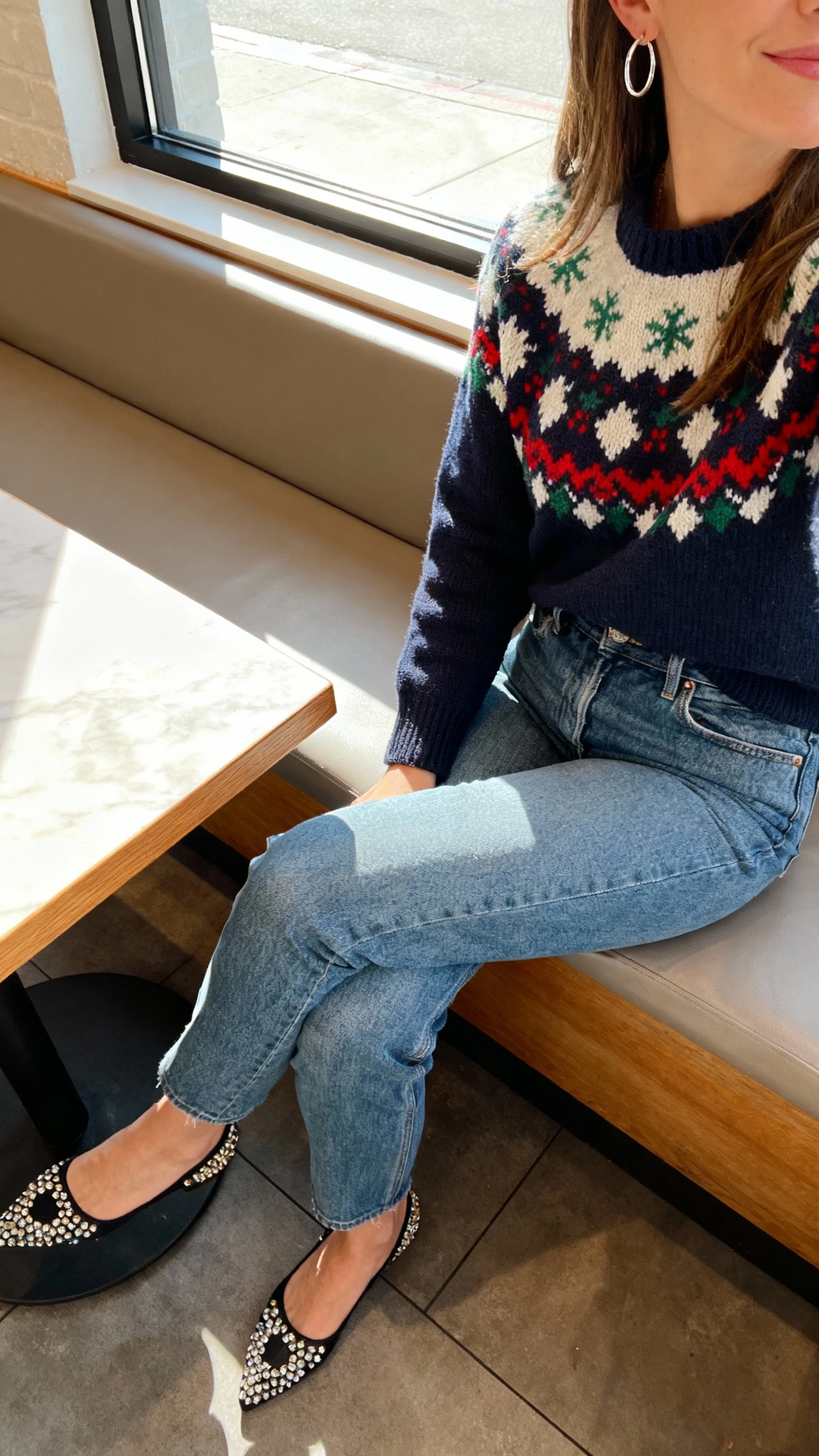 iPhone cafe table selfie cropped at shoulders showing a woman in elevated jeans with a festive knit sweater and embellished flats, simple hoop earrings, face not visible, window seat daylight, natural iPhone aesthetic.