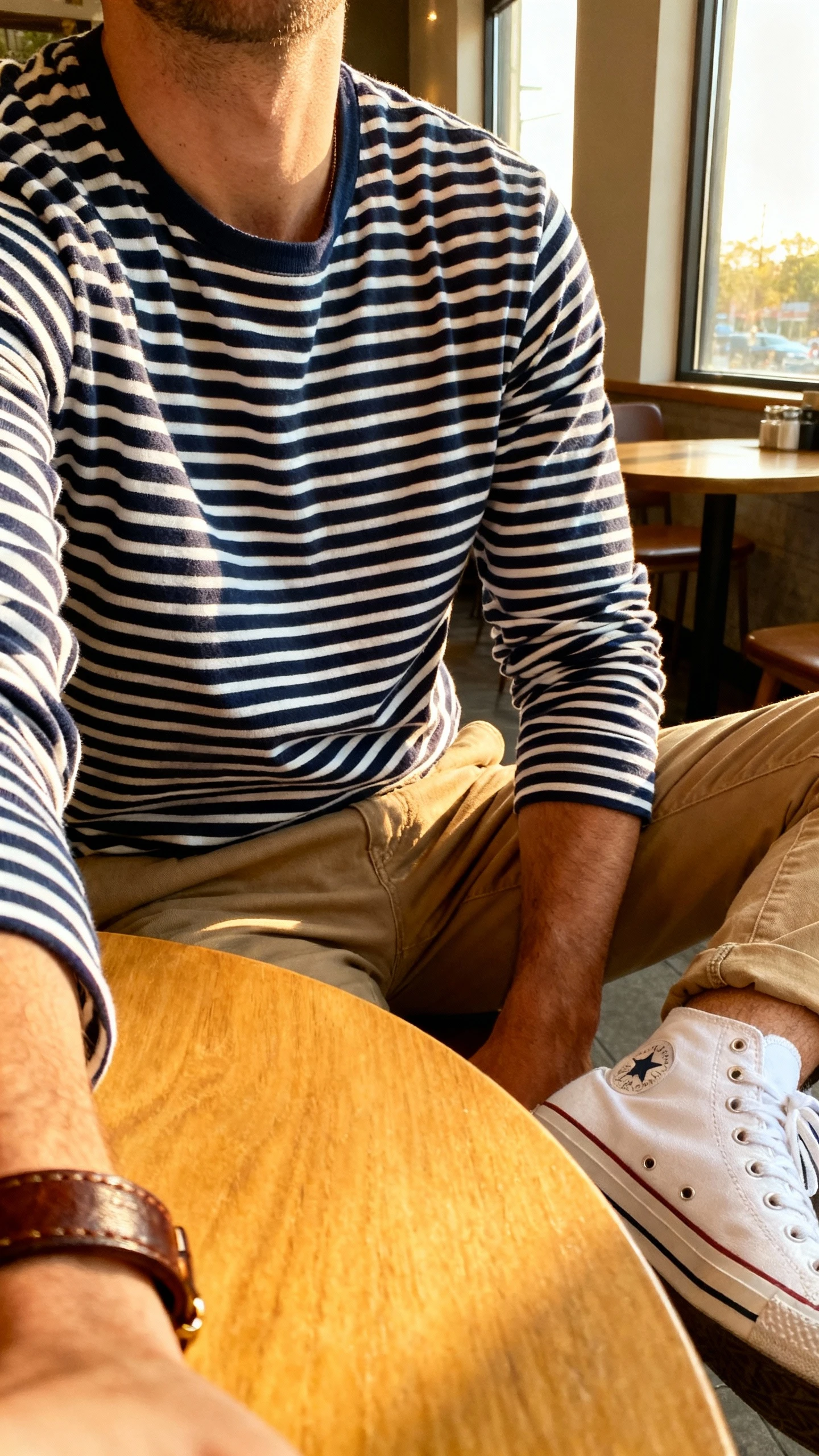 iPhone cafe table selfie cropped at shoulders showing a man wearing a navy-and-white Breton stripe long-sleeve tee with classic khakis, white canvas sneakers, and a leather bracelet, face not visible, warm afternoon window light, natural iPhone aesthetic.