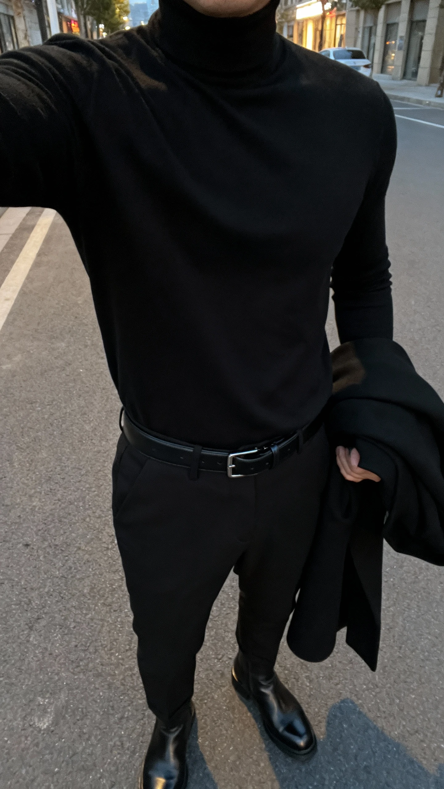 iPhone chest-down selfie of a man in an all-black outfit: black turtleneck, black tailored trousers, black leather belt, sleek black boots, and a minimalist black overcoat draped over arm, face not visible, city street at dusk, natural ambient light, iPhone photo quality.