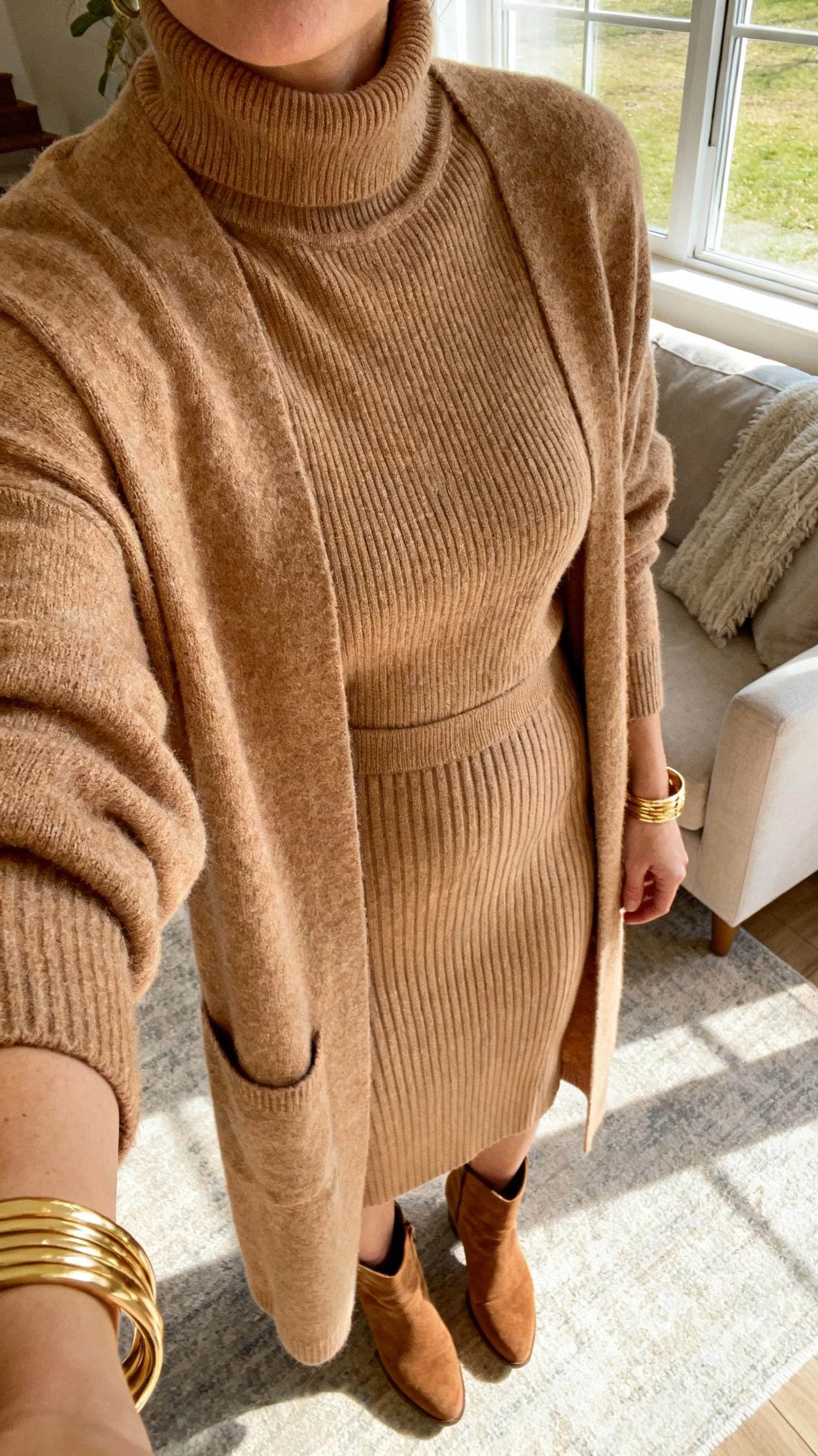 iPhone close-crop selfie at shoulders of a woman in head-to-toe tonal knit layers: camel turtleneck, matching longline cardigan, and ribbed knit skirt, suede ankle boots, gold bangles, face not visible, living room by window, warm afternoon light, natural iPhone photo quality.