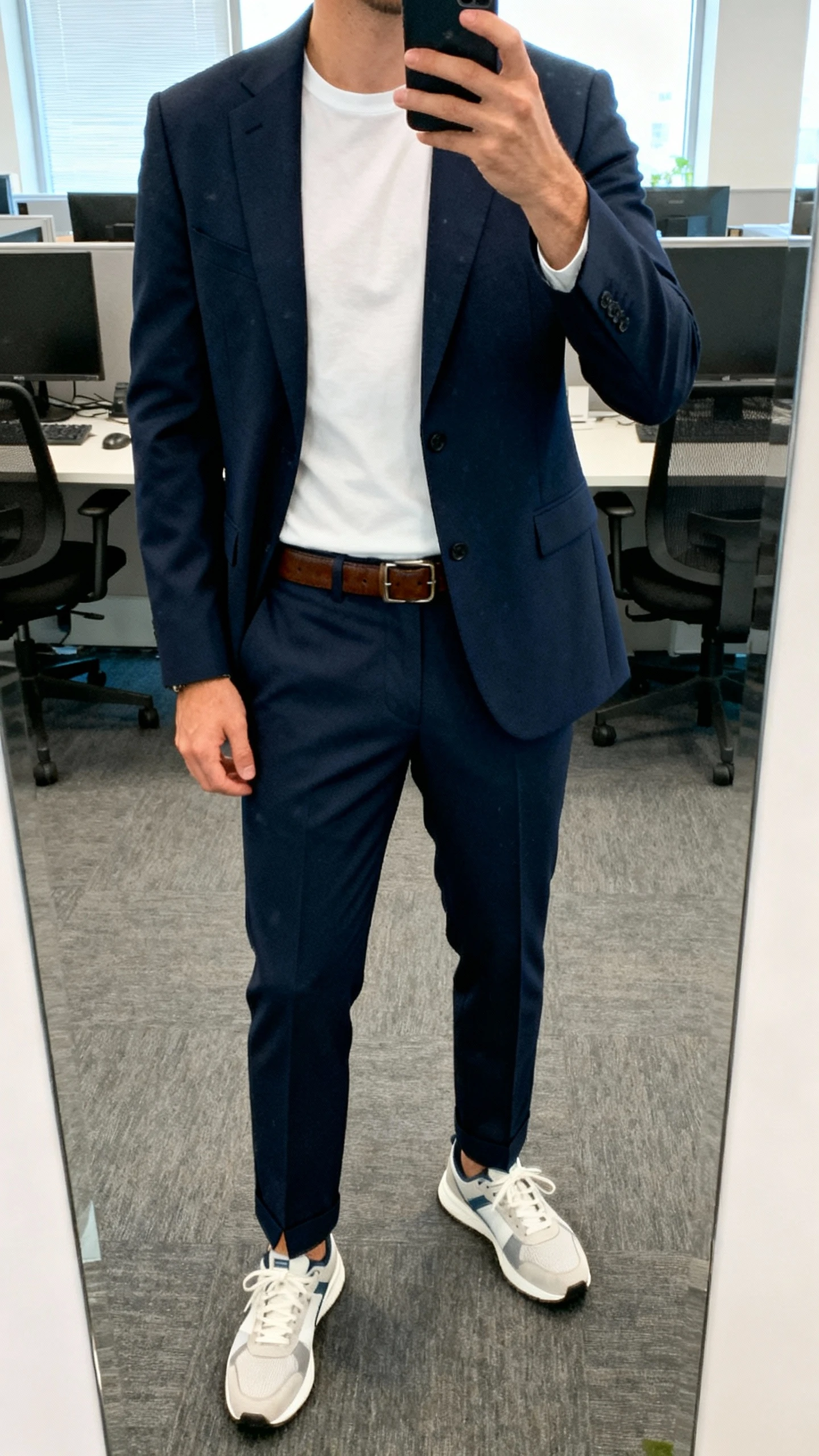 iPhone cropped-at-neck office mirror selfie of a man wearing a navy suit jacket with a white tee and the matching suit trousers split up with clean sneakers, leather belt, face not visible, bright office light, iPhone photo.