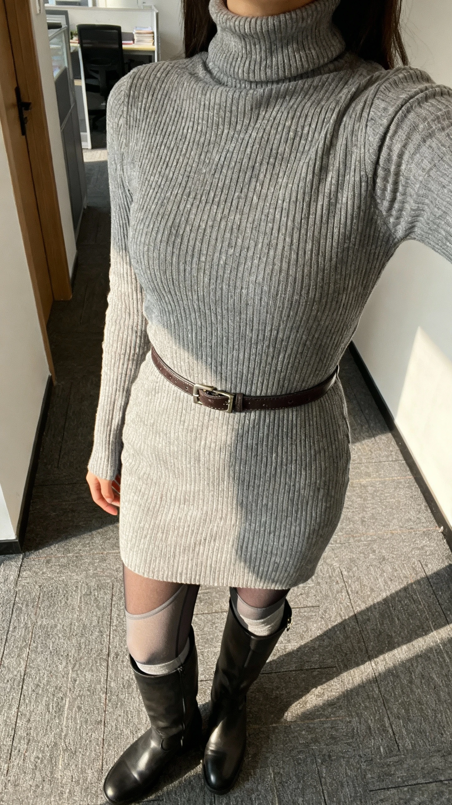 iPhone cropped-at-shoulders selfie of a woman wearing a ribbed turtleneck dress, knee-high boots, and subtle thermal tights underneath, belt at waist, face not visible, office corridor with morning light, natural iPhone photo quality.