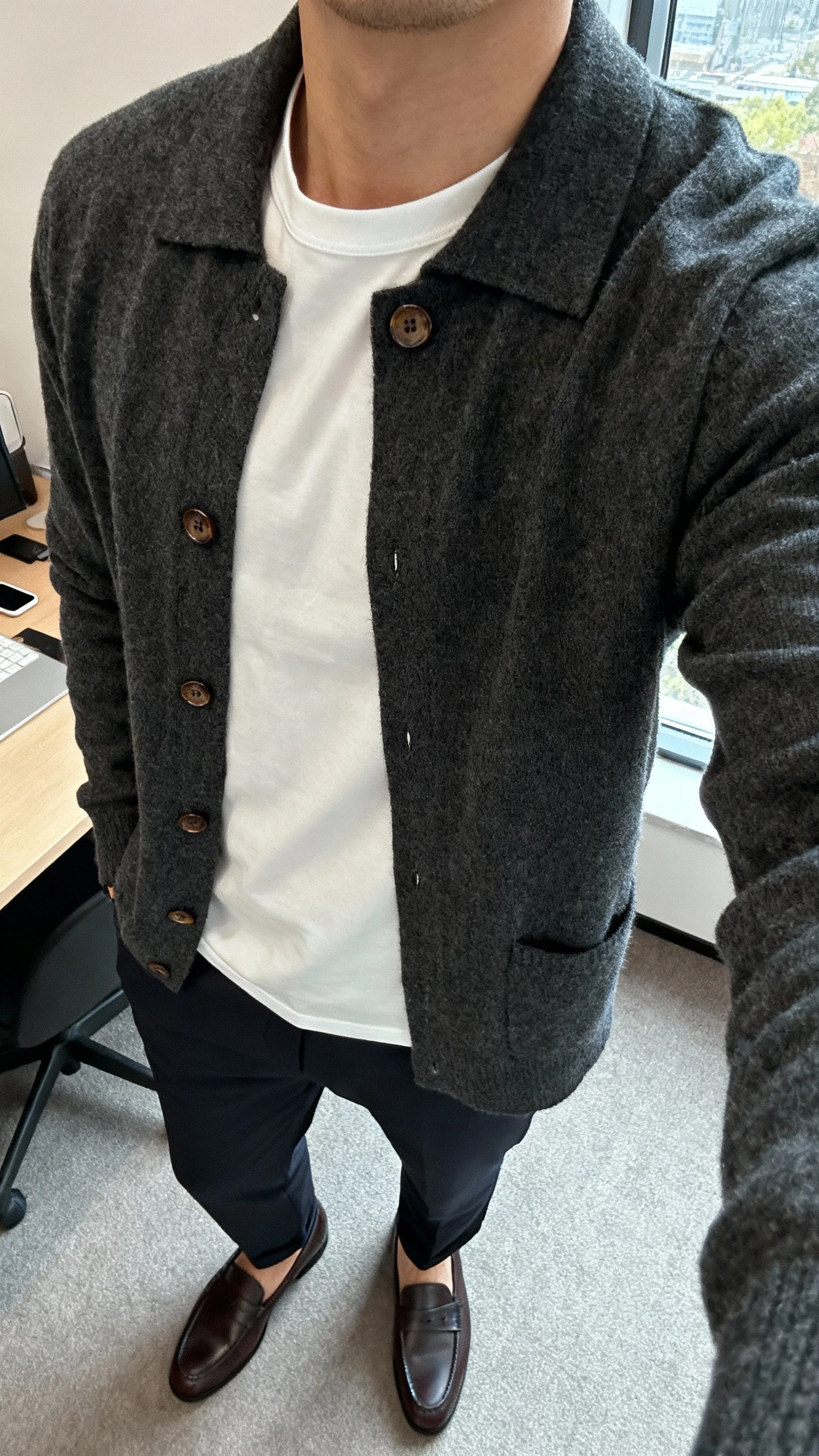 iPhone desk selfie cropped at neck showing a man wearing a buttoned charcoal cardigan over a crisp white tee with dark chinos and loafers, face not visible, daylight from window, natural iPhone photo quality.