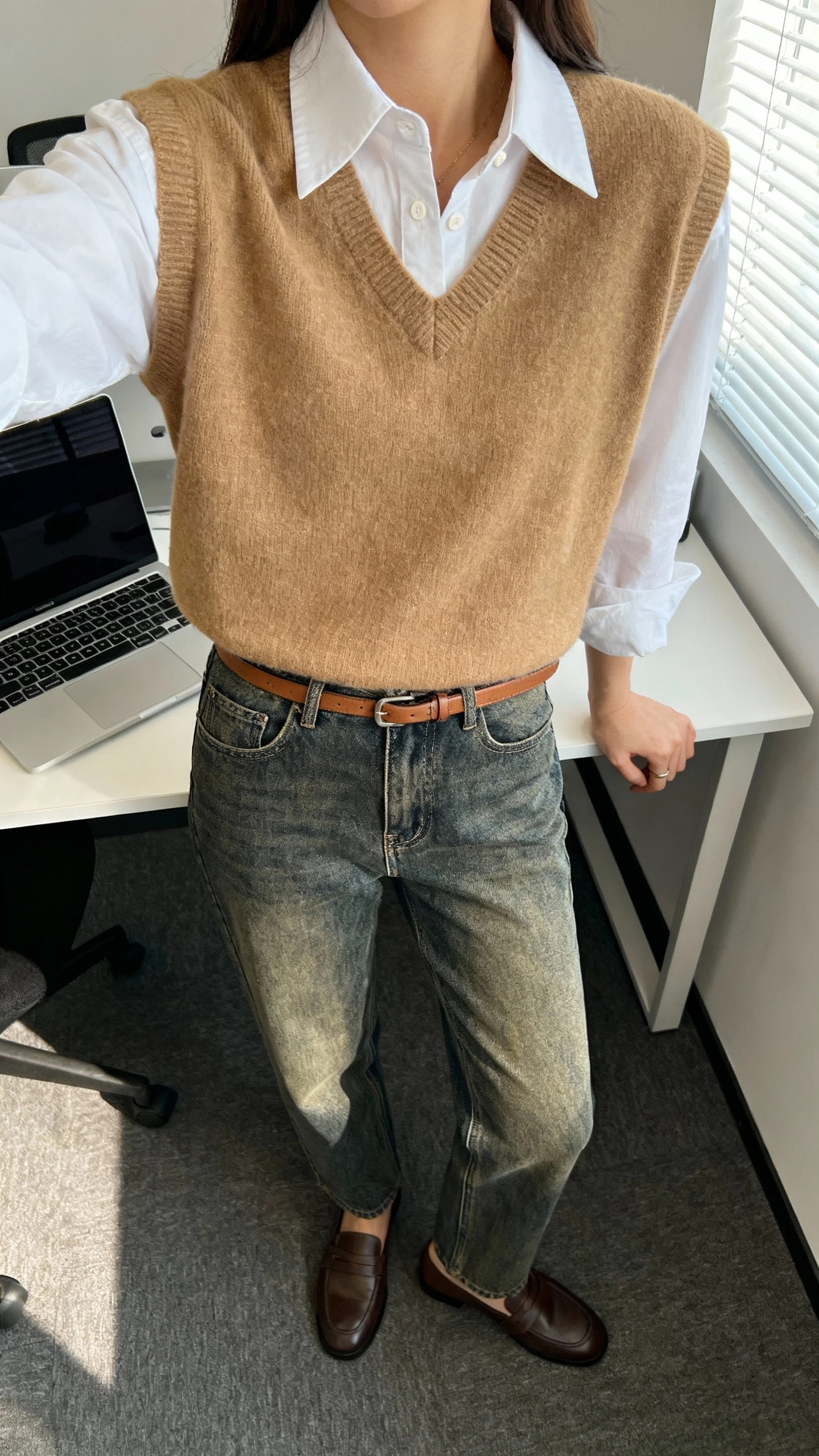 iPhone desk selfie cropped at neck showing a woman in a crisp white button-down layered with a camel sweater vest, mid-wash straight jeans, loafers, and a thin leather belt, face not visible, office setting with laptop, natural window light, casual iPhone photo.