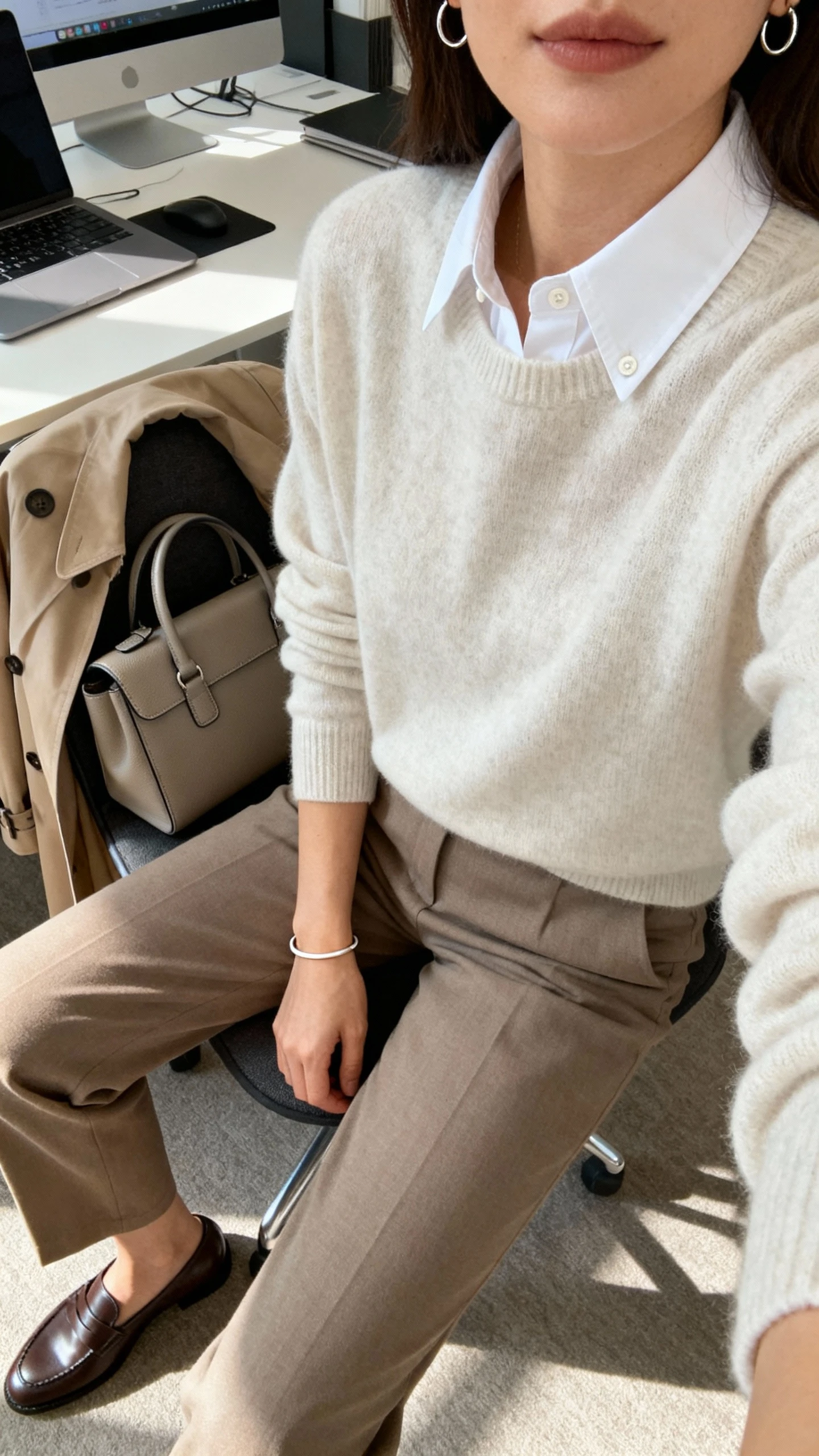 iPhone desk selfie cropped at shoulders of a woman layering a crisp white button-up under a crewneck sweater, straight-leg trousers, loafers, minimal bracelet, small hoops, structured satchel on chair, optional trench draped over back; face not visible, office morning light.