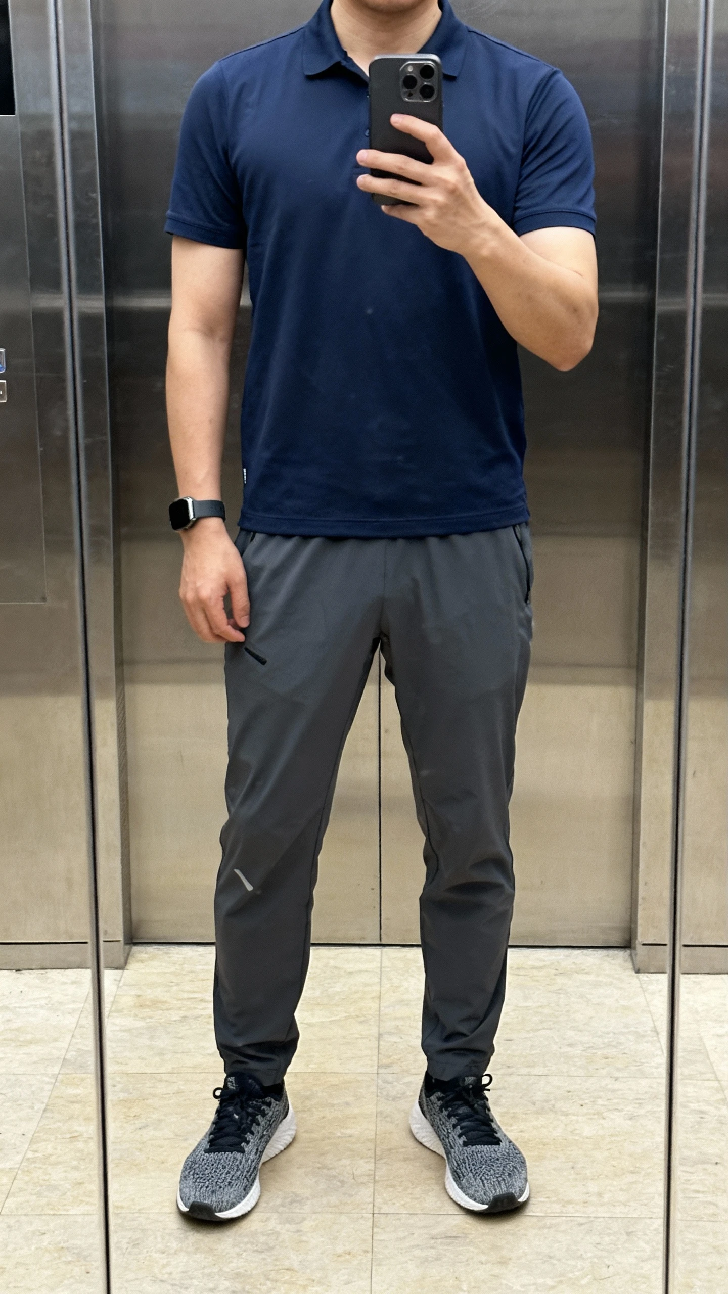 iPhone elevator mirror selfie of a man wearing a navy polo shirt with charcoal tech pants, knit sneakers, and a fitness watch, face not visible, natural indoor lighting, everyday iPhone photo quality.