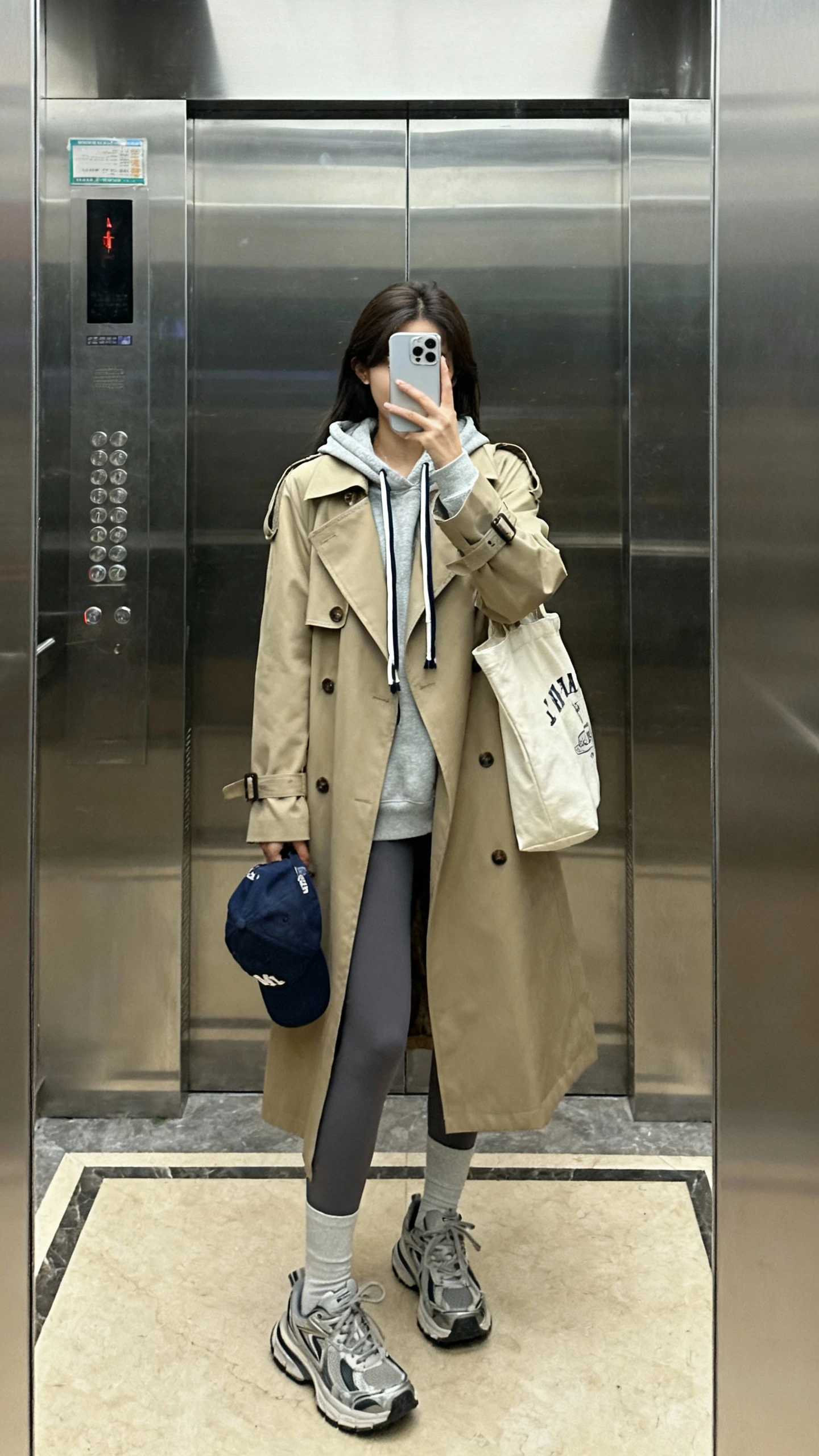 iPhone elevator mirror selfie of a woman in a classic trench coat layered over a hoodie with drawstrings showing, leggings, chunky sneakers, baseball cap tucked under arm, tote bag, face not visible, indoor lighting, natural iPhone aesthetic.