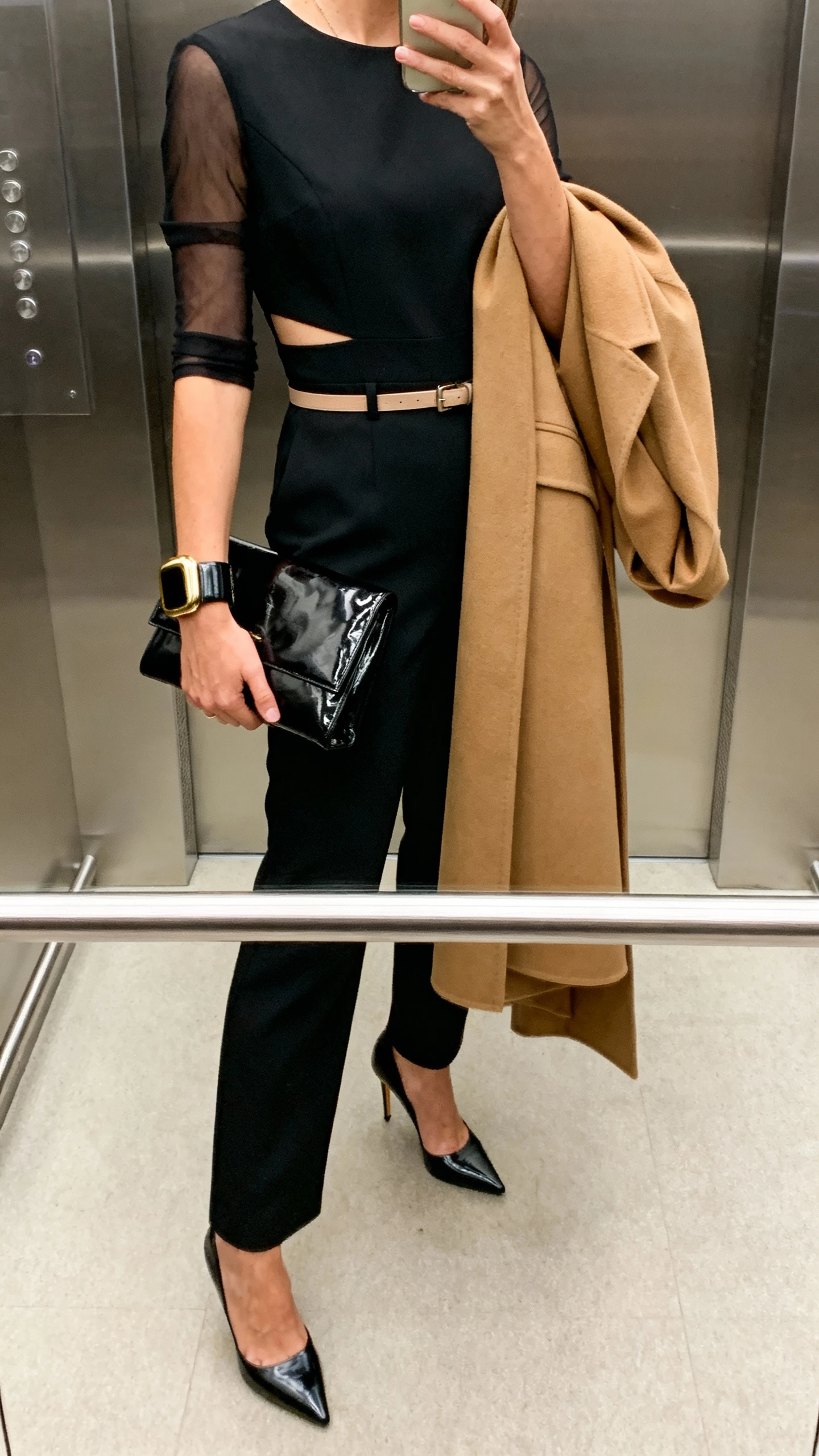 iPhone elevator mirror selfie of a woman in a tailored black jumpsuit with sheer sleeves and subtle waist cutouts, sculptural heels, patent leather clutch, bold cuff bracelet, thin waist belt, and a longline camel coat draped over the arm; face not visible, indoor lighting, casual iPhone photo.