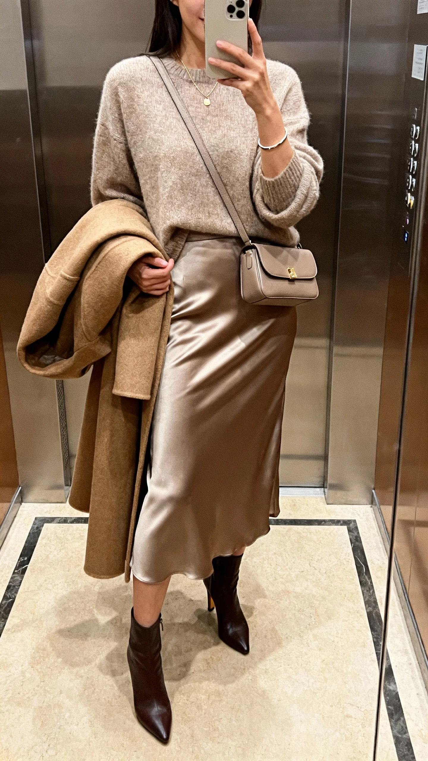 iPhone elevator mirror selfie of a woman wearing a midi satin bias-cut skirt with a relaxed cashmere sweater, heeled ankle boots, delicate necklace, slim bangle, mini shoulder bag, classic wool coat over arm; face not visible, indoor lighting, casual iPhone shot.