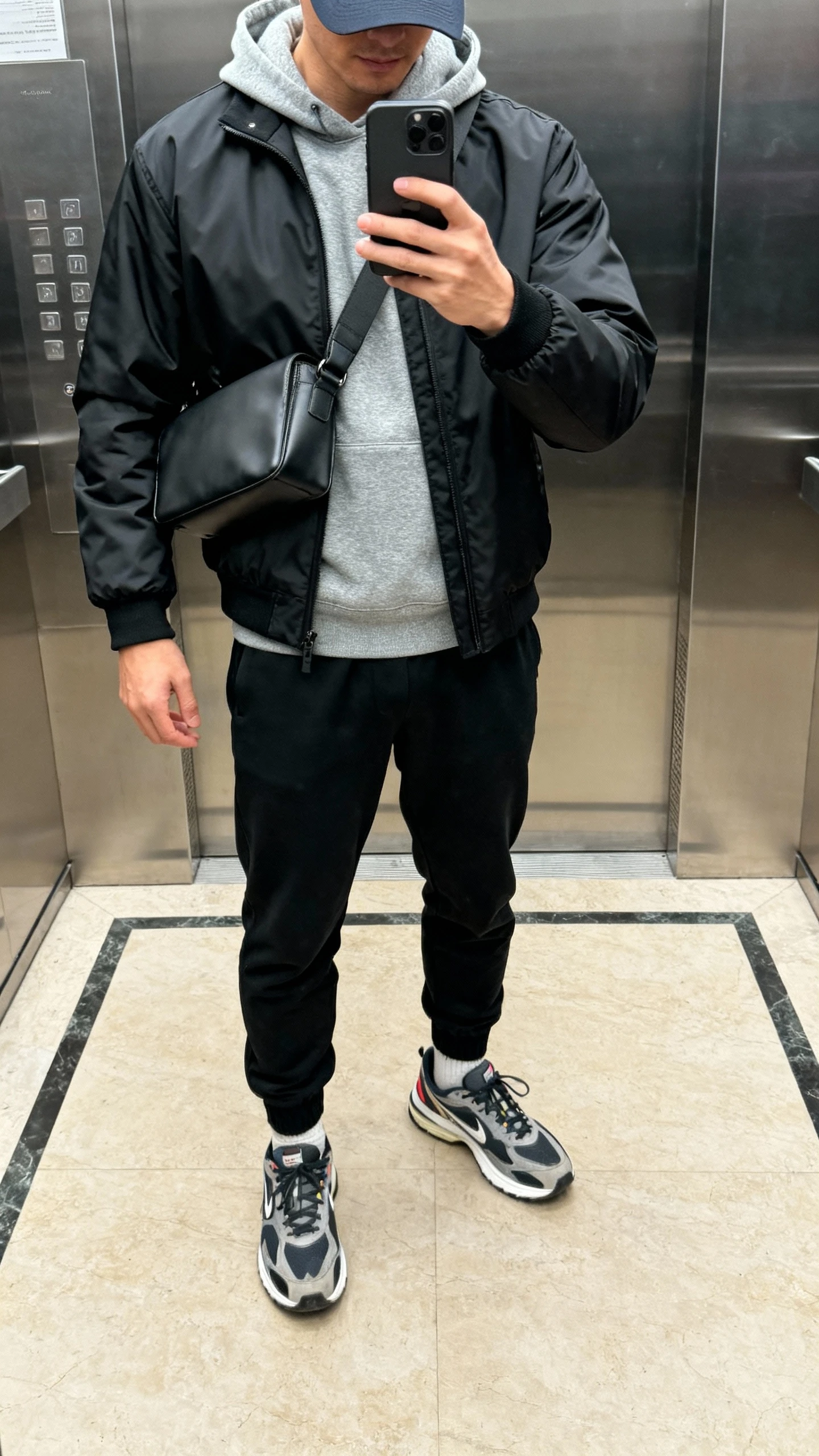 iPhone elevator selfie cropped at shoulders showing a man in a gray premium hoodie under a matte black technical shell, tapered black joggers, retro running sneakers, a sleek crossbody bag, and a tonal cap; indoor lighting, face not visible, natural iPhone photo.