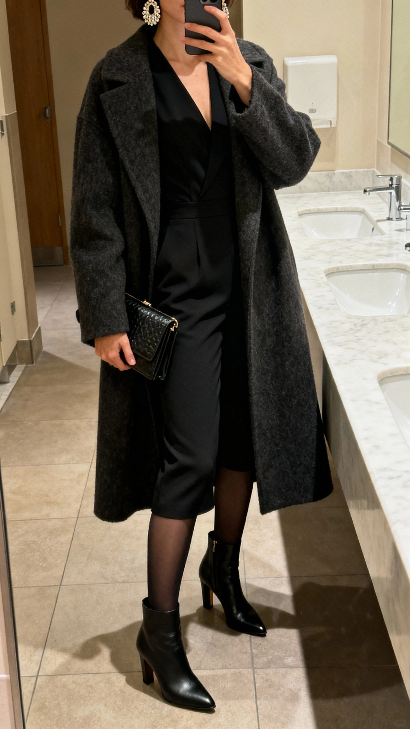 iPhone evening restroom mirror selfie of a woman wearing a tailored black jumpsuit with heeled ankle boots, long wool coat, opaque tights underneath, structured clutch, statement earrings; face not visible, warm indoor lighting, natural iPhone photo quality.