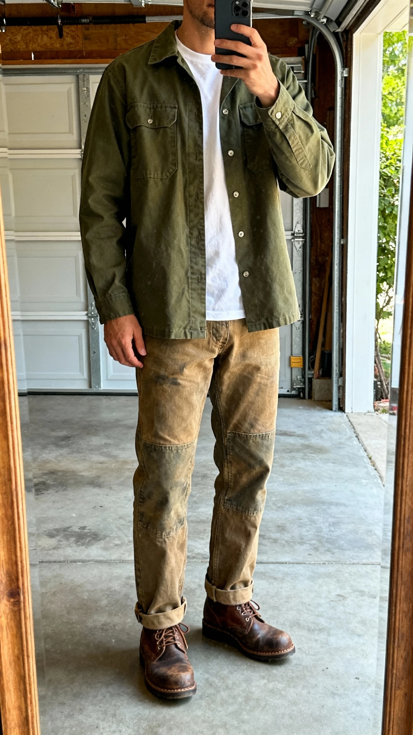 iPhone garage mirror selfie of a man wearing an olive workwear-inspired overshirt over a white tee with rugged canvas pants and moc-toe boots, face not visible, cool natural light, casual iPhone quality.