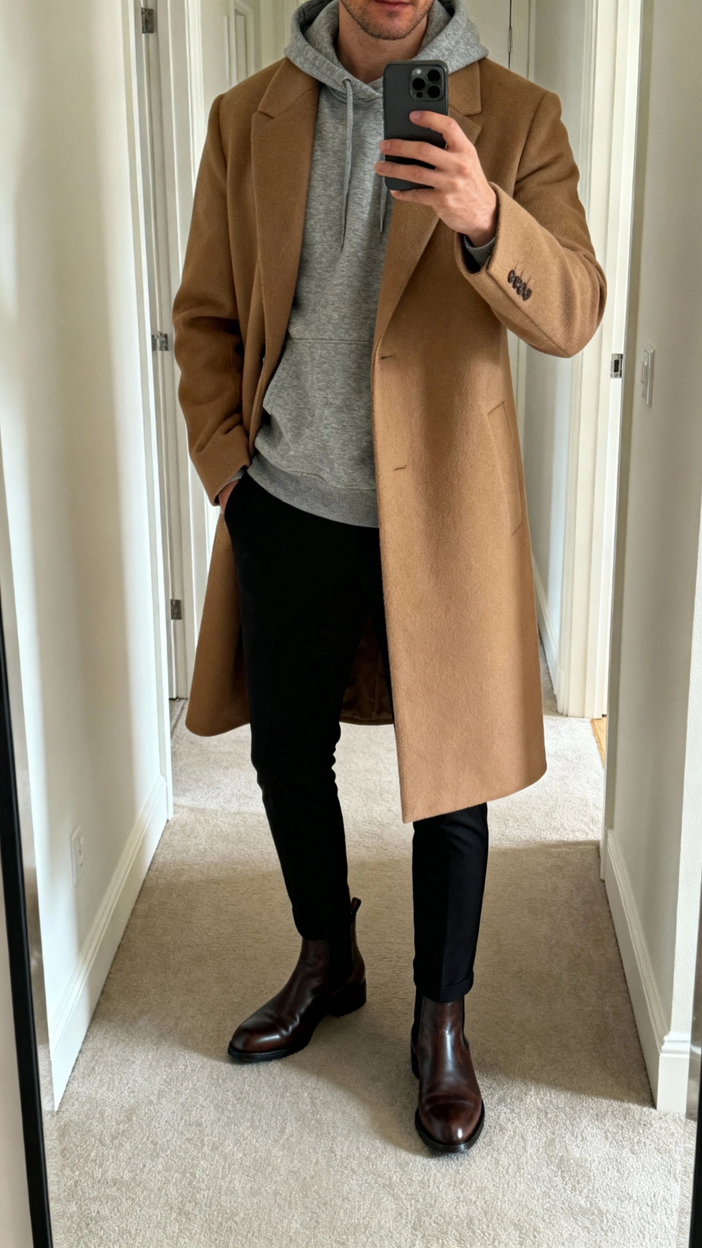 iPhone hallway mirror selfie of a man wearing a heather gray hoodie layered under a camel overcoat with slim black trousers and leather Chelsea boots, face not visible, soft indoor lighting, natural iPhone quality.