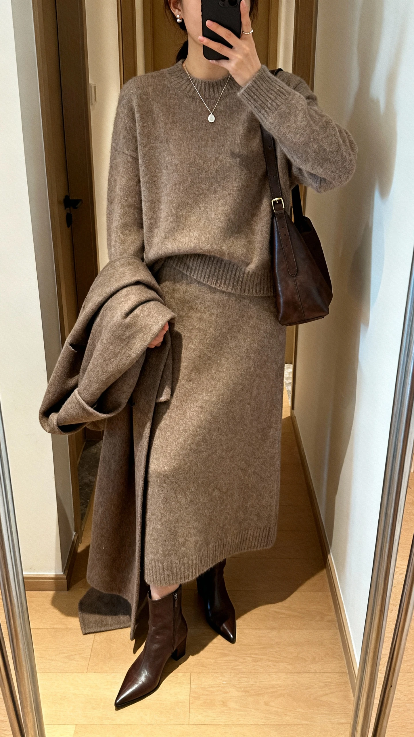 iPhone hallway mirror selfie of a woman wearing a matching wool-cashmere sweater and knit skirt set, pointed ankle boots, longline wool coat draped over arm, delicate pendant necklace, small stud earrings, leather shoulder bag; face not visible, natural indoor lighting, casual iPhone aesthetic.