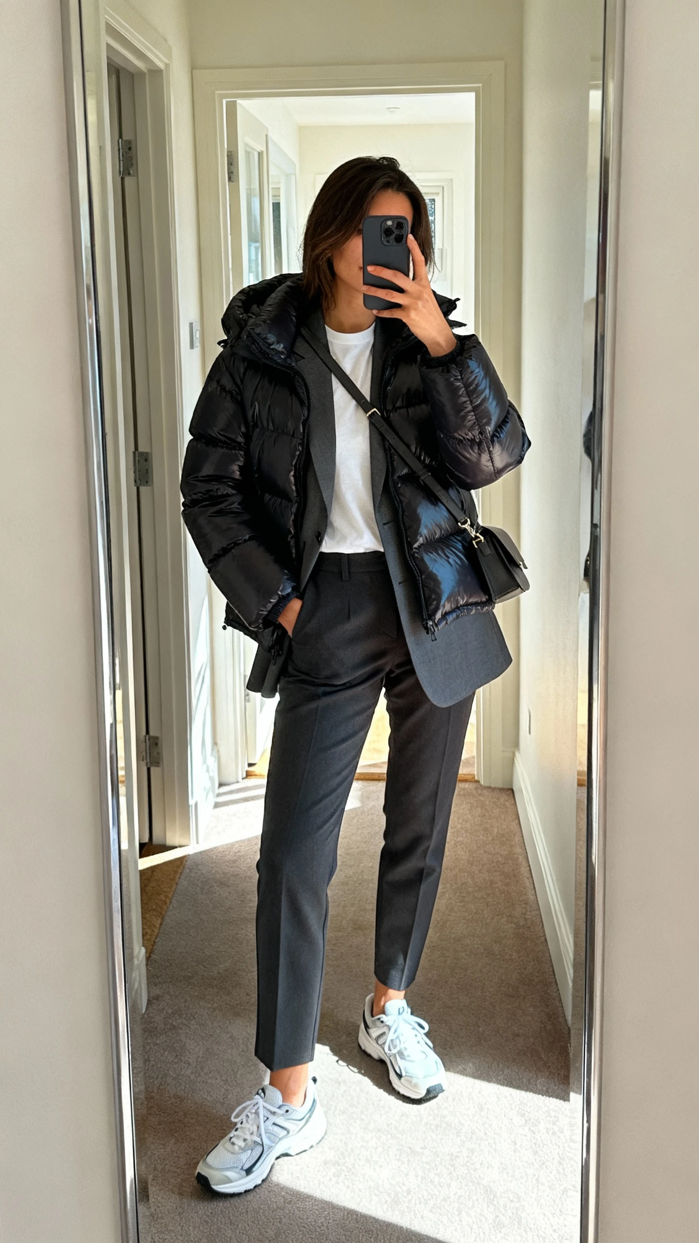 iPhone hallway mirror selfie of a woman wearing a sleek puffer jacket layered over a tailored blazer with a tee underneath, tapered trousers, clean sneakers, crossbody bag, face not visible, apartment entry mirror, afternoon daylight, casual iPhone photo.