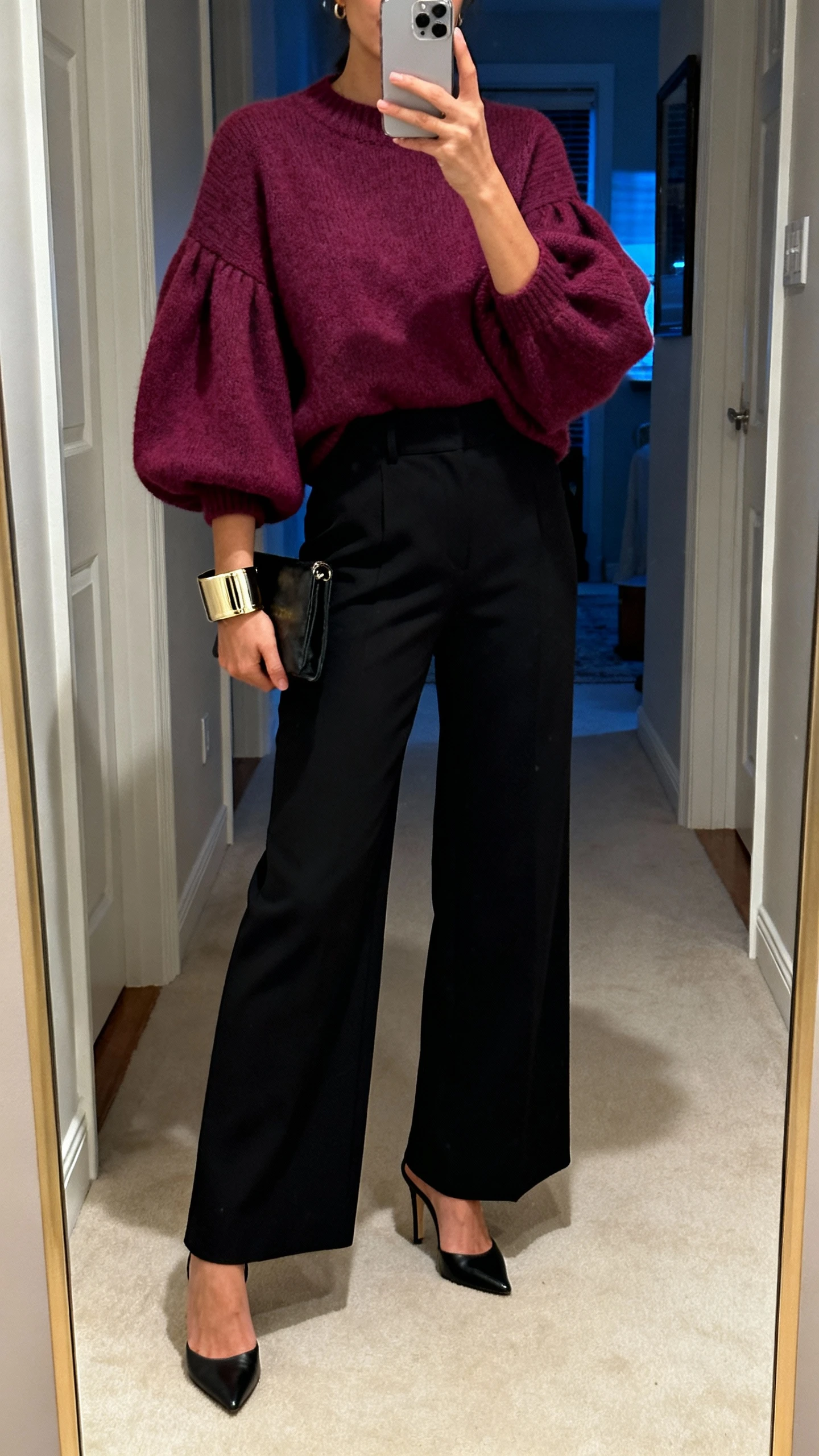 iPhone hallway mirror selfie of a woman wearing a statement sleeve sweater (balloon sleeves in berry) with high-waist wide-leg trousers (black), heeled mules, bold cuff bracelet, small clutch, face not visible, evening indoor lighting, natural iPhone aesthetic.