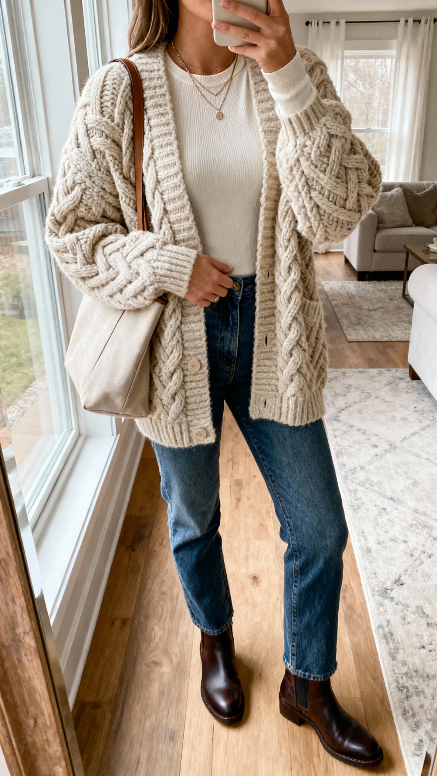 iPhone living room mirror selfie of a woman in a chunky cable-knit statement cardigan over a fitted long-sleeve base layer, straight-leg jeans, Chelsea boots, casual tote, subtle layered necklaces; face not visible, natural window light, casual iPhone photo.