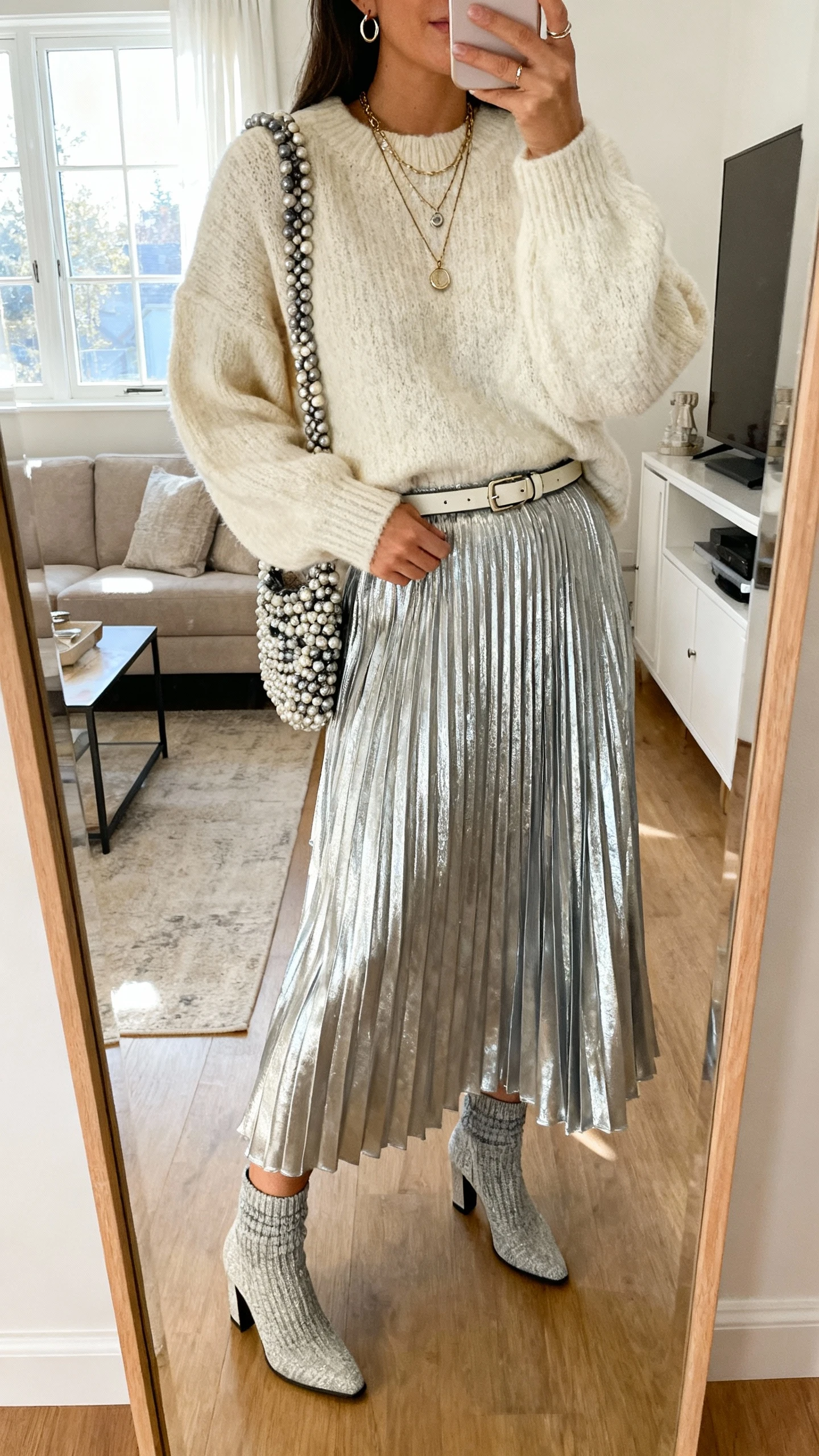 iPhone living-room mirror selfie of a woman wearing a silver metallic pleated maxi skirt, chunky cream sweater, heeled sock boots, beaded shoulder bag, layered pendant necklaces, small hoops, and a slim waist-cinching belt; face not visible, morning window light, natural iPhone photo quality.