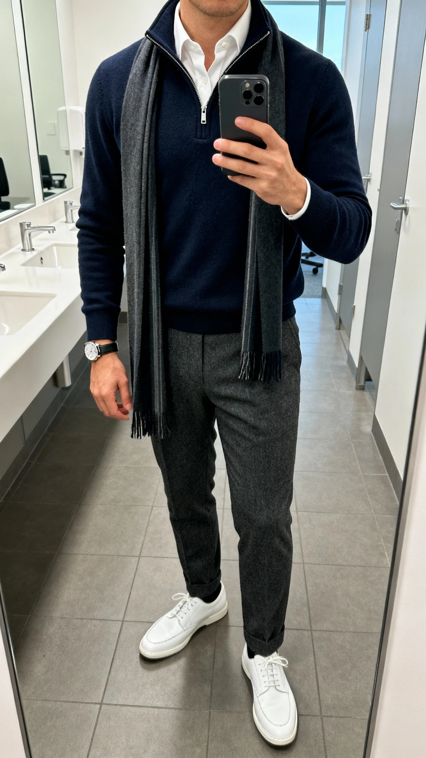 iPhone mirror selfie cropped at neck showing a man wearing a navy merino half-zip over a white Oxford shirt, charcoal wool trousers, white leather sneakers, a slim scarf draped, and a simple watch; office restroom mirror, bright indoor light, face not visible, natural iPhone quality.