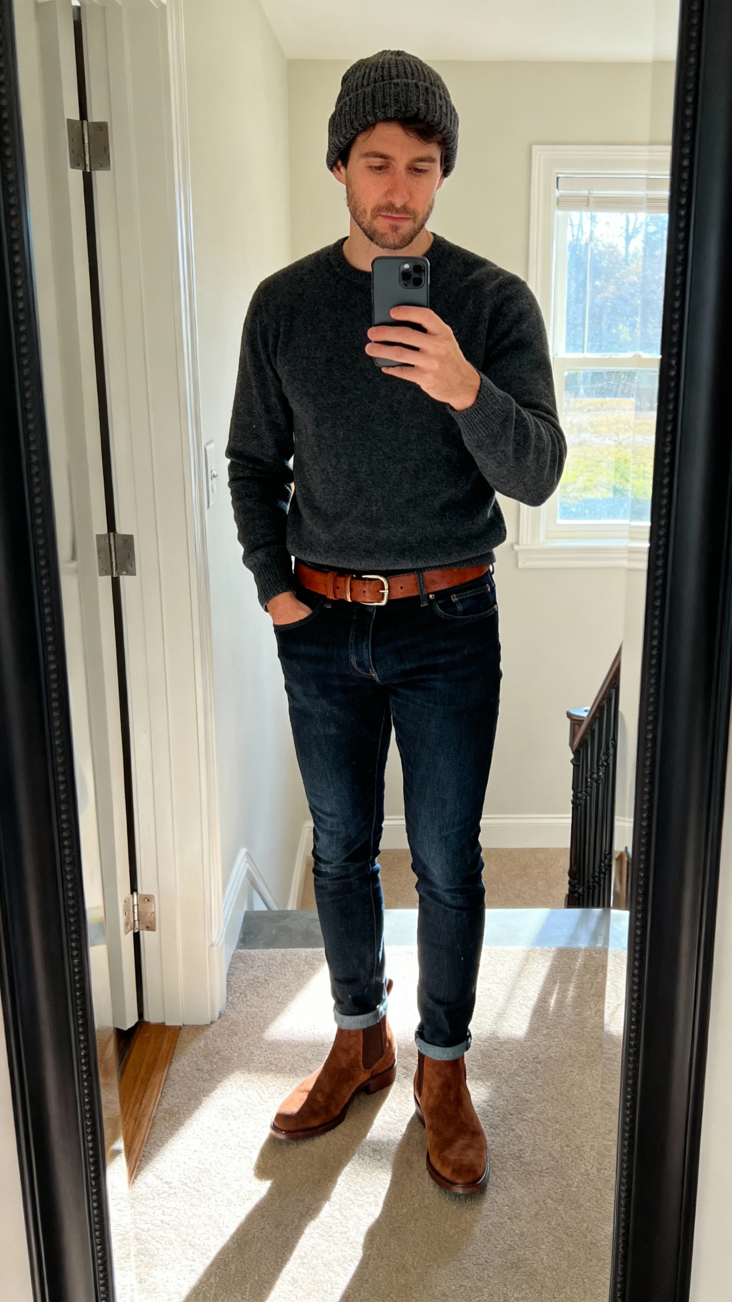iPhone mirror selfie cropped at shoulders showing a man in dark indigo slim jeans, a charcoal merino crewneck sweater, brown suede Chelsea boots, matching brown leather belt, and a wool beanie; entryway mirror, afternoon window light, face not visible, iPhone photo quality.