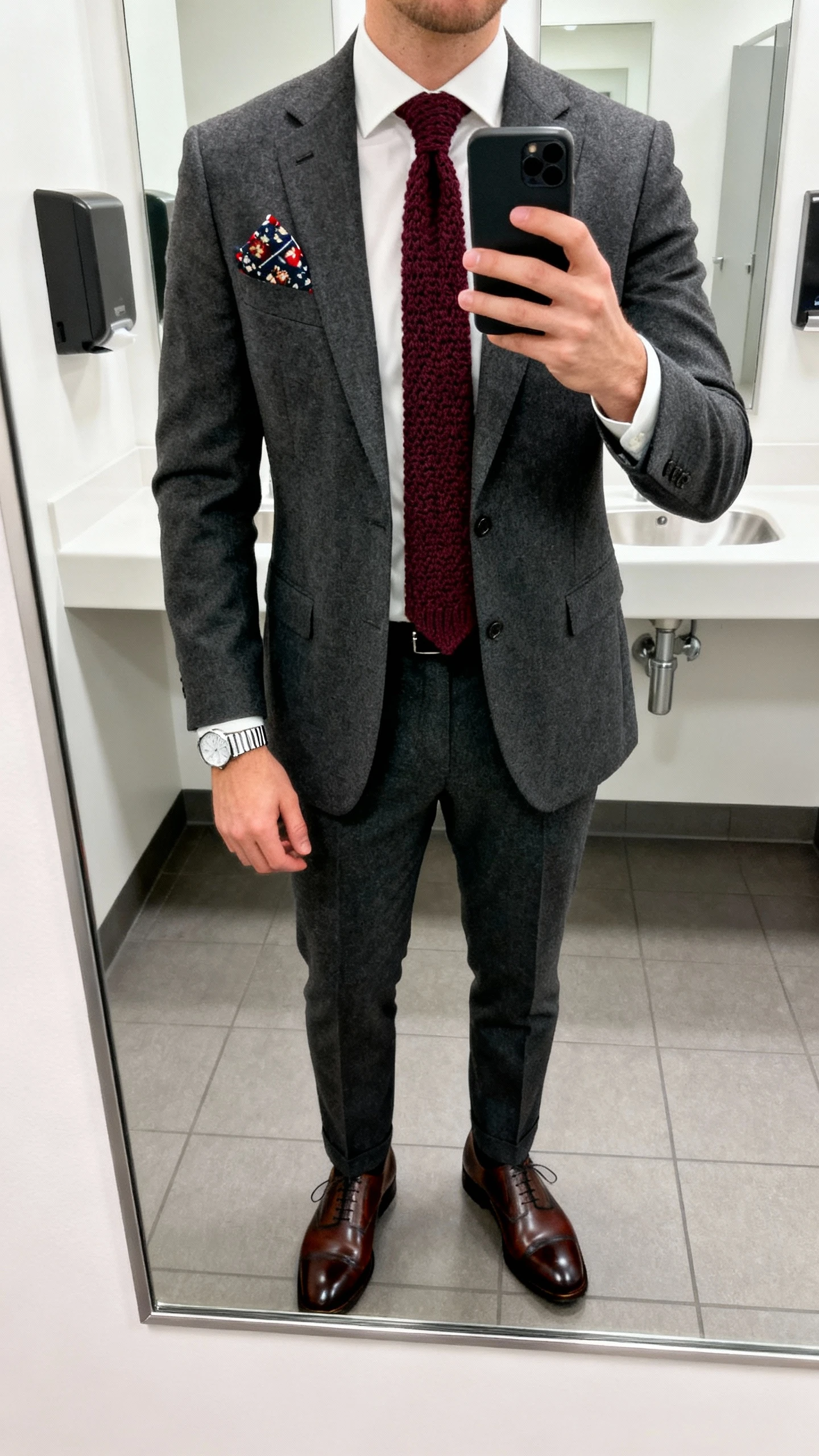 iPhone mirror selfie of a man in a classic suit with a festive twist: charcoal suit, white dress shirt, burgundy knit tie, patterned pocket square, polished oxford shoes, and a slim silver watch, office restroom mirror under bright indoor light, face not visible, natural iPhone photo quality.
