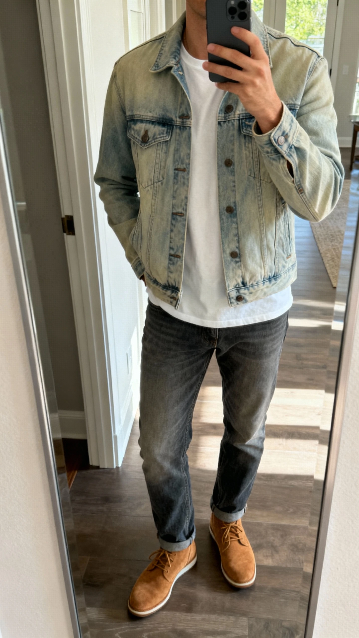 iPhone mirror selfie of a man in denim-on-denim: light-wash trucker jacket over a white tee with mid-wash jeans and tan suede sneakers, face not visible, entryway mirror with daylight, casual iPhone photo.