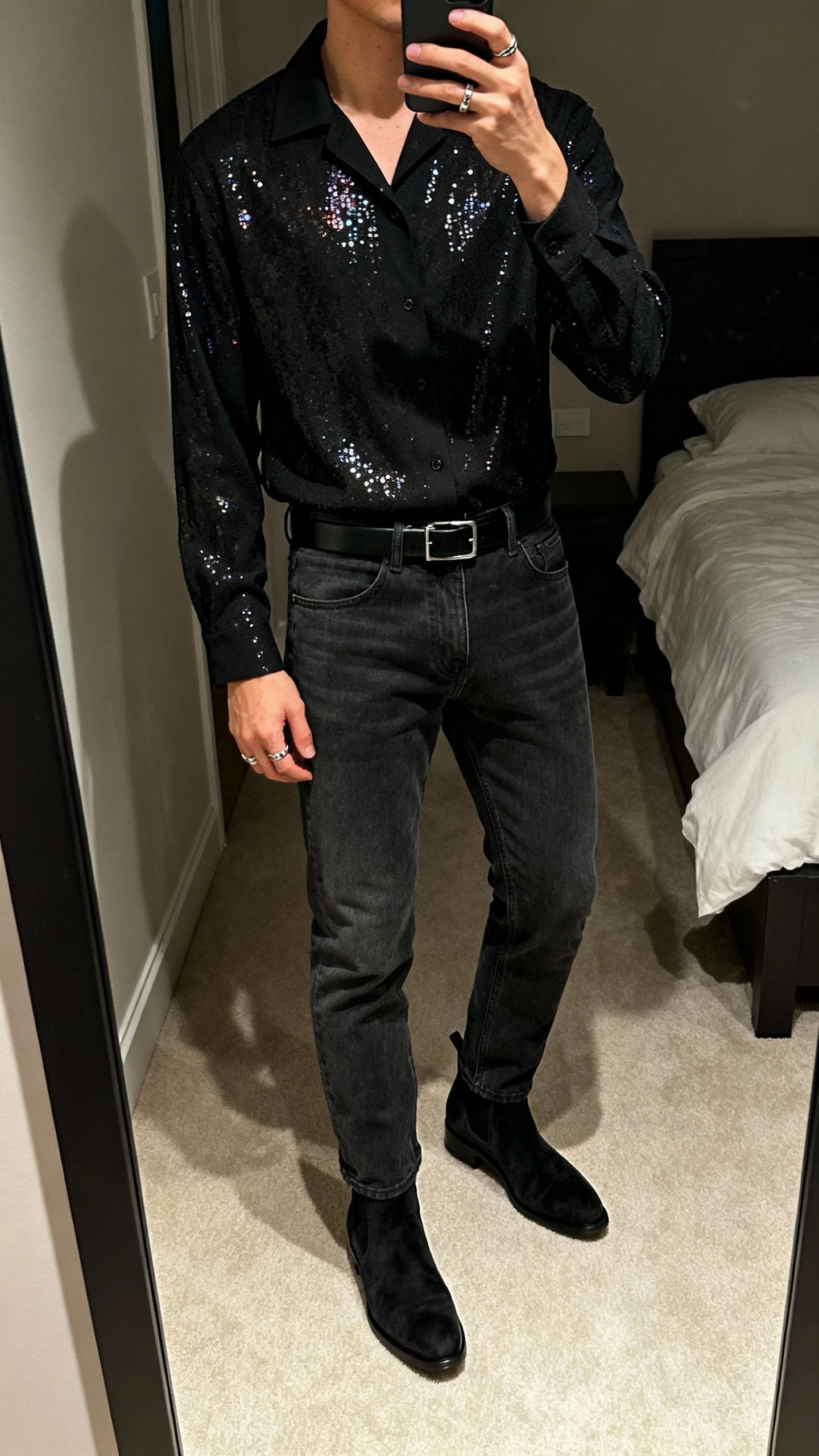 iPhone mirror selfie of a man wearing a black camp-collar shirt with subtle tonal sequin detailing, dark charcoal jeans, black suede Chelsea boots, slim black belt, minimal rings, face not visible, bedroom mirror, evening indoor lighting, natural iPhone photo quality