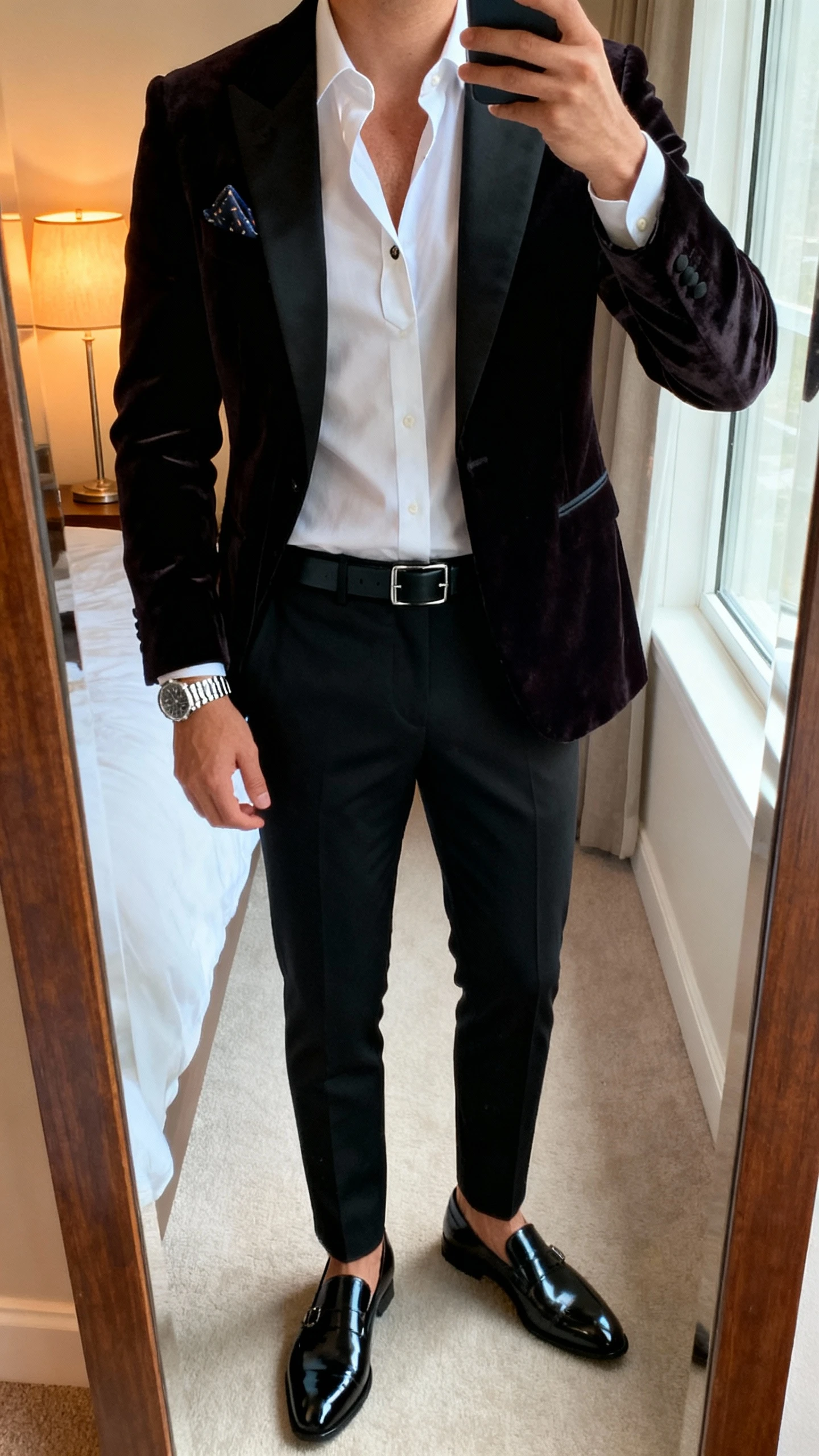 iPhone mirror selfie of a man wearing a dark midnight velvet dinner jacket, crisp white dress shirt with undone top button, slim black trousers, black patent loafers, slim black belt, silver watch, pocket square, face not visible, cropped at neck, bedroom mirror with warm lamp and window light, natural iPhone photo quality