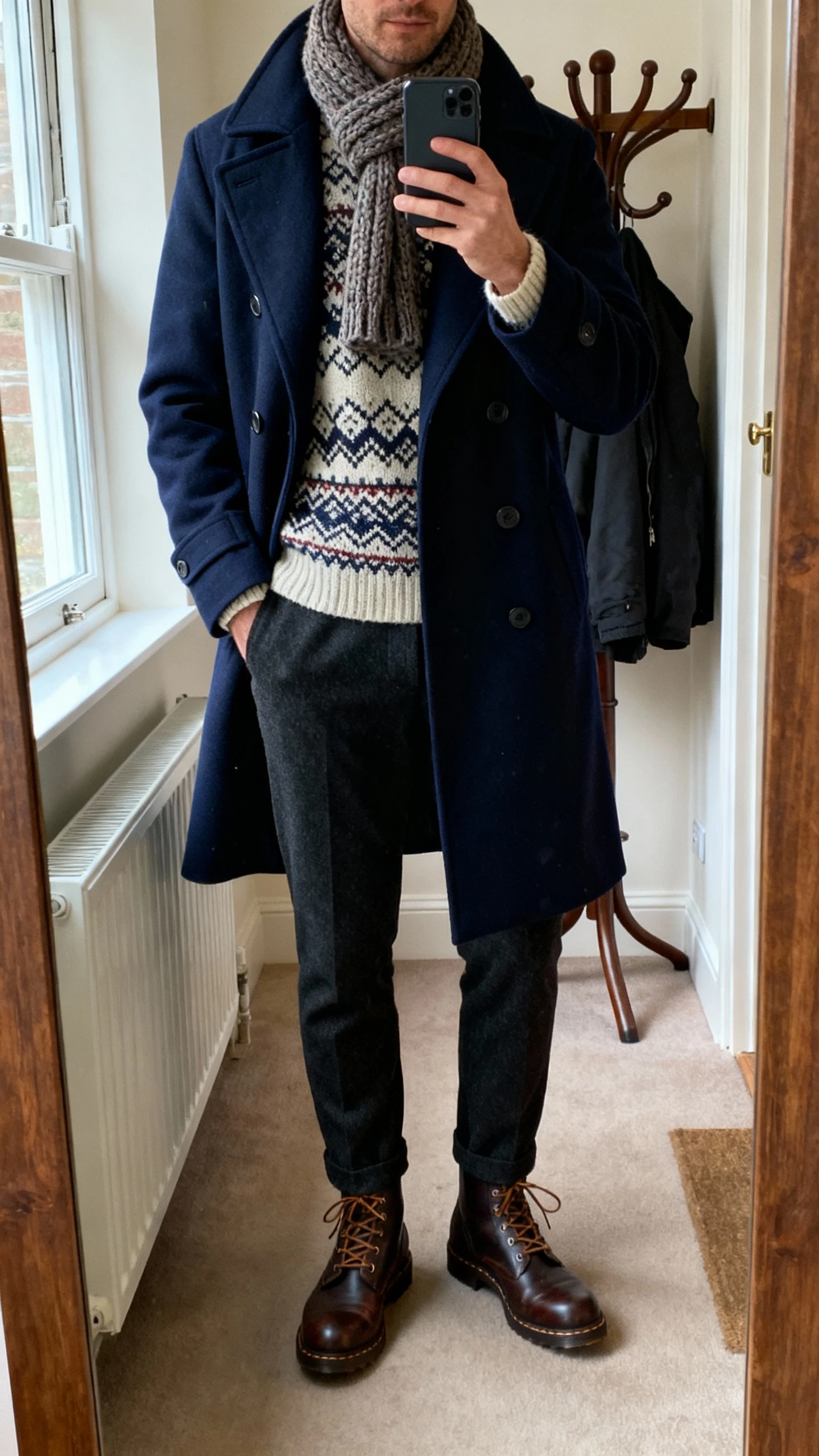 iPhone mirror selfie of a man wearing a navy peacoat over a Nordic patterned knit sweater, dark wool trousers, lace-up boots, and a knit scarf; entryway mirror with coat rack, morning window light, cropped at neck, face not visible, natural iPhone photo.