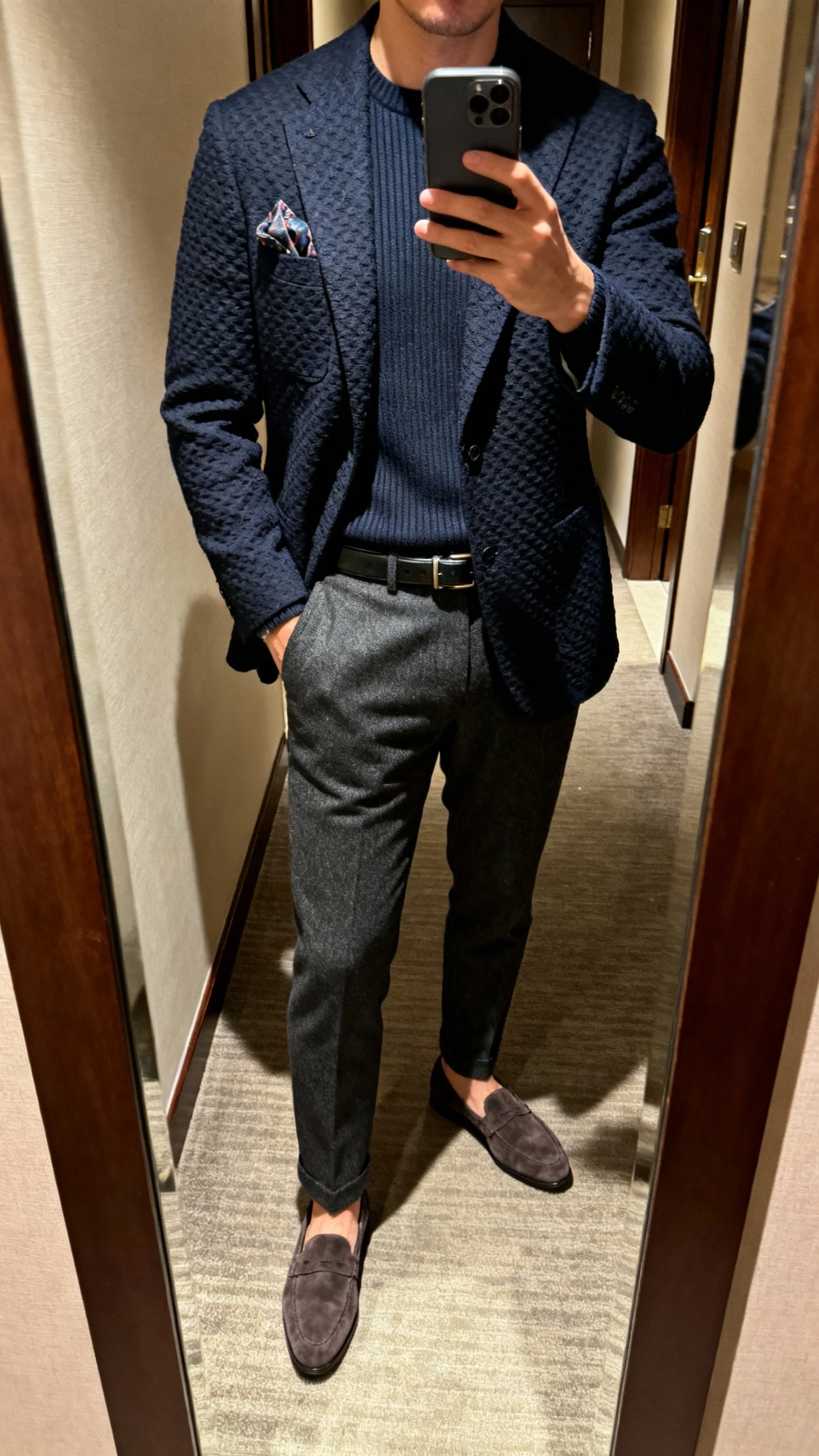 iPhone mirror selfie of a man wearing a navy textured blazer over a fine-gauge crewneck knit, charcoal wool trousers, suede loafers, slim leather belt, and a pocket square; hotel hallway mirror, soft evening lighting, cropped at neck, face not visible, natural iPhone photo.