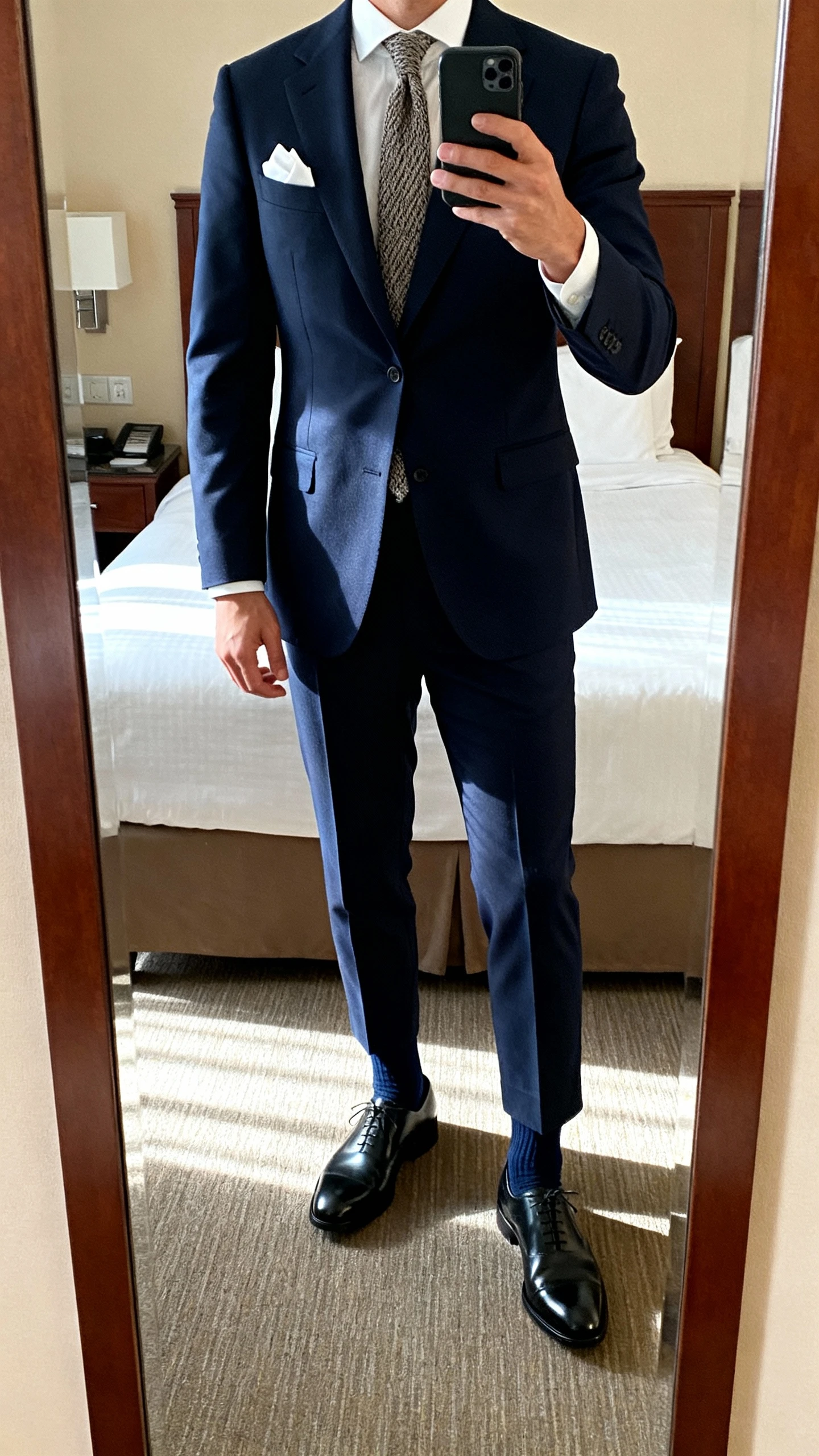 iPhone mirror selfie of a man wearing a navy two-button suit, white Oxford shirt, textured knit silk tie, black cap-toe oxfords, white pocket square, and navy dress socks; hotel room full-length mirror, morning light, face not visible, natural iPhone quality.