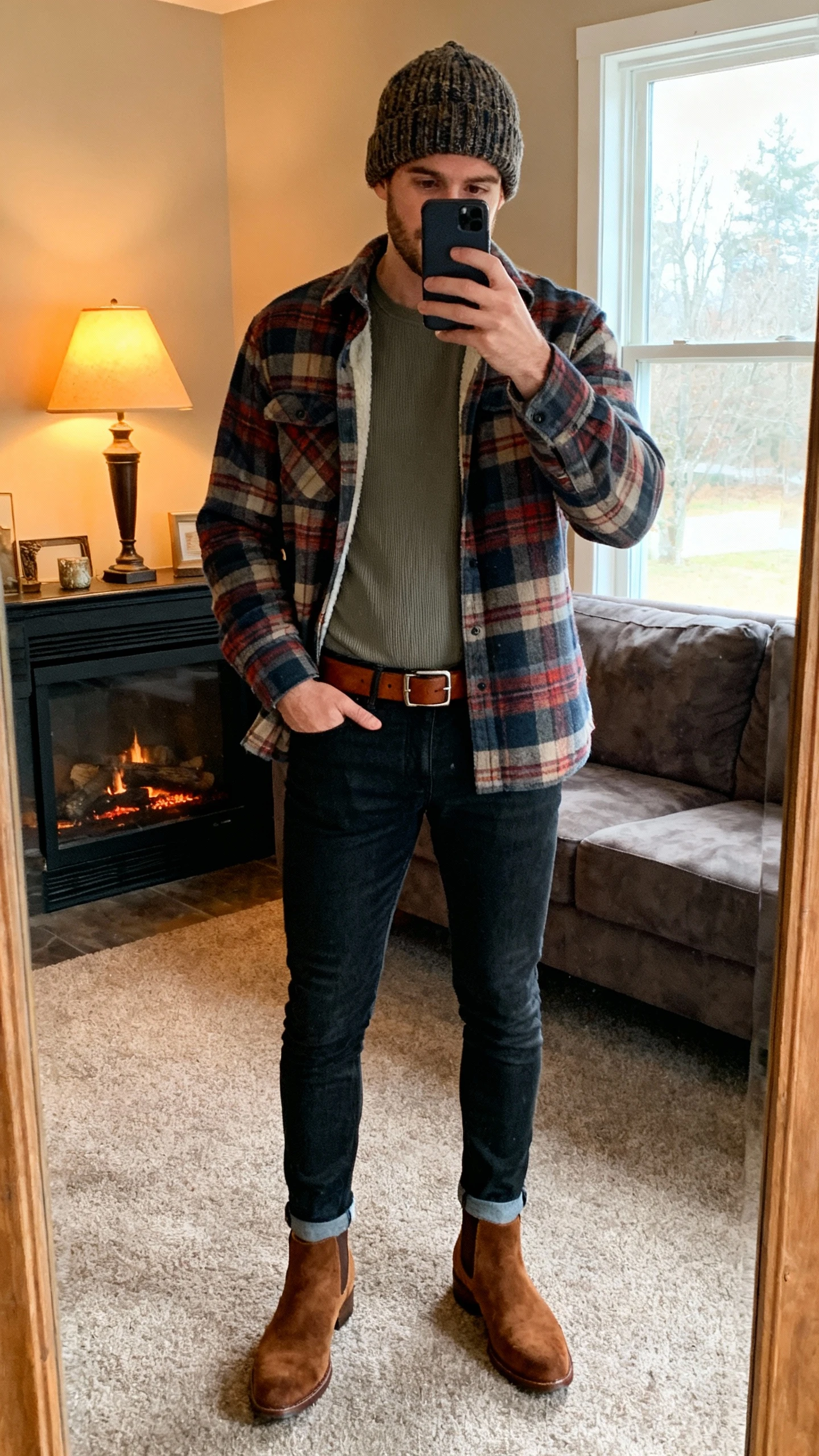 iPhone mirror selfie of a man wearing a plaid flannel shirt layered over a thermal tee, slim dark jeans, brown suede Chelsea boots, leather belt, and a knit beanie, face not visible, cropped at neck, cozy living room by a fireplace, warm indoor lamp and window light, natural iPhone photo quality.