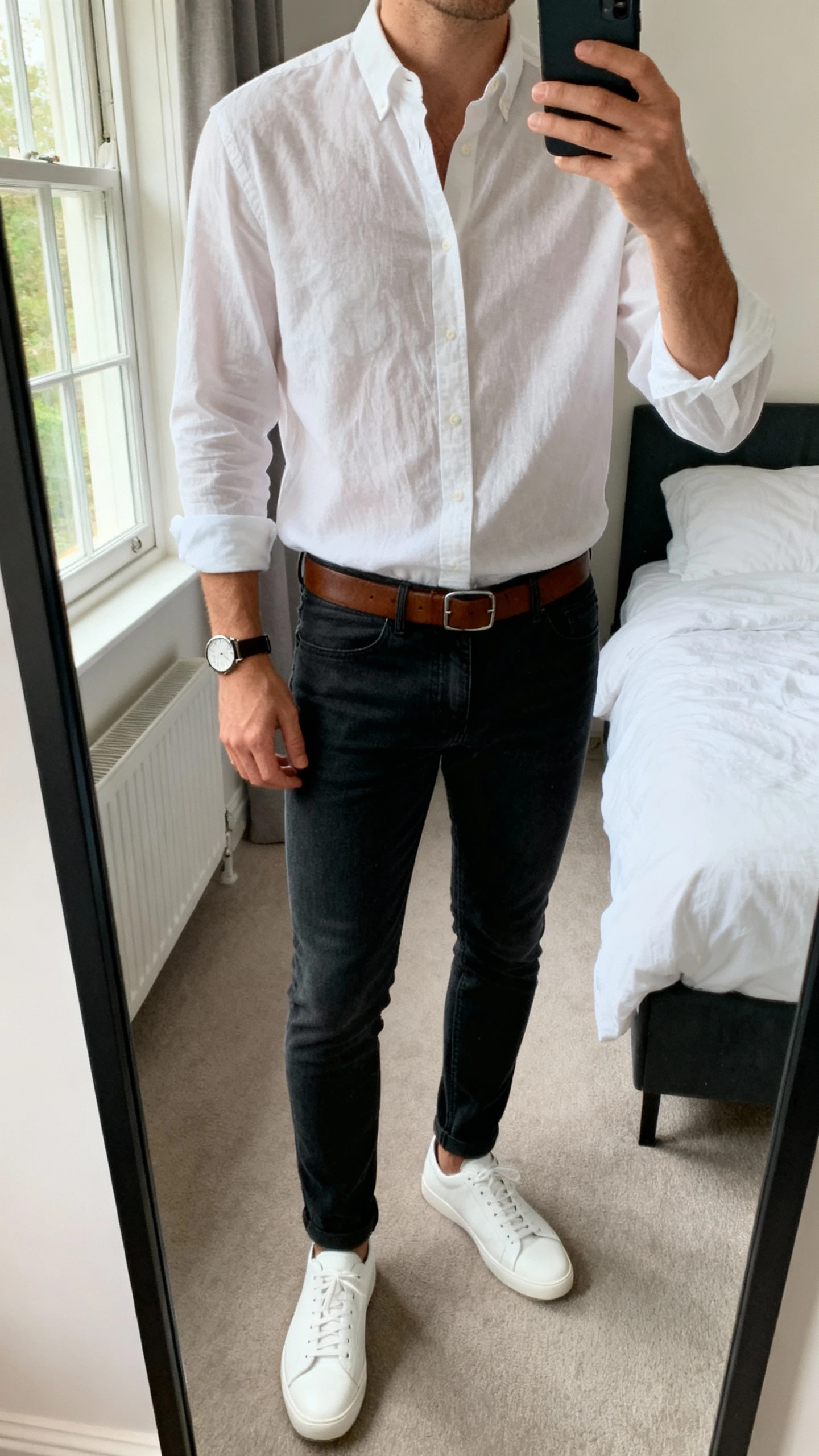 iPhone mirror selfie of a man wearing a white soft-wash Oxford shirt tucked into dark slim jeans, white leather sneakers, brown leather belt, and a simple everyday watch; cropped at neck, bedroom mirror, natural window light, face not visible, natural iPhone photo quality.