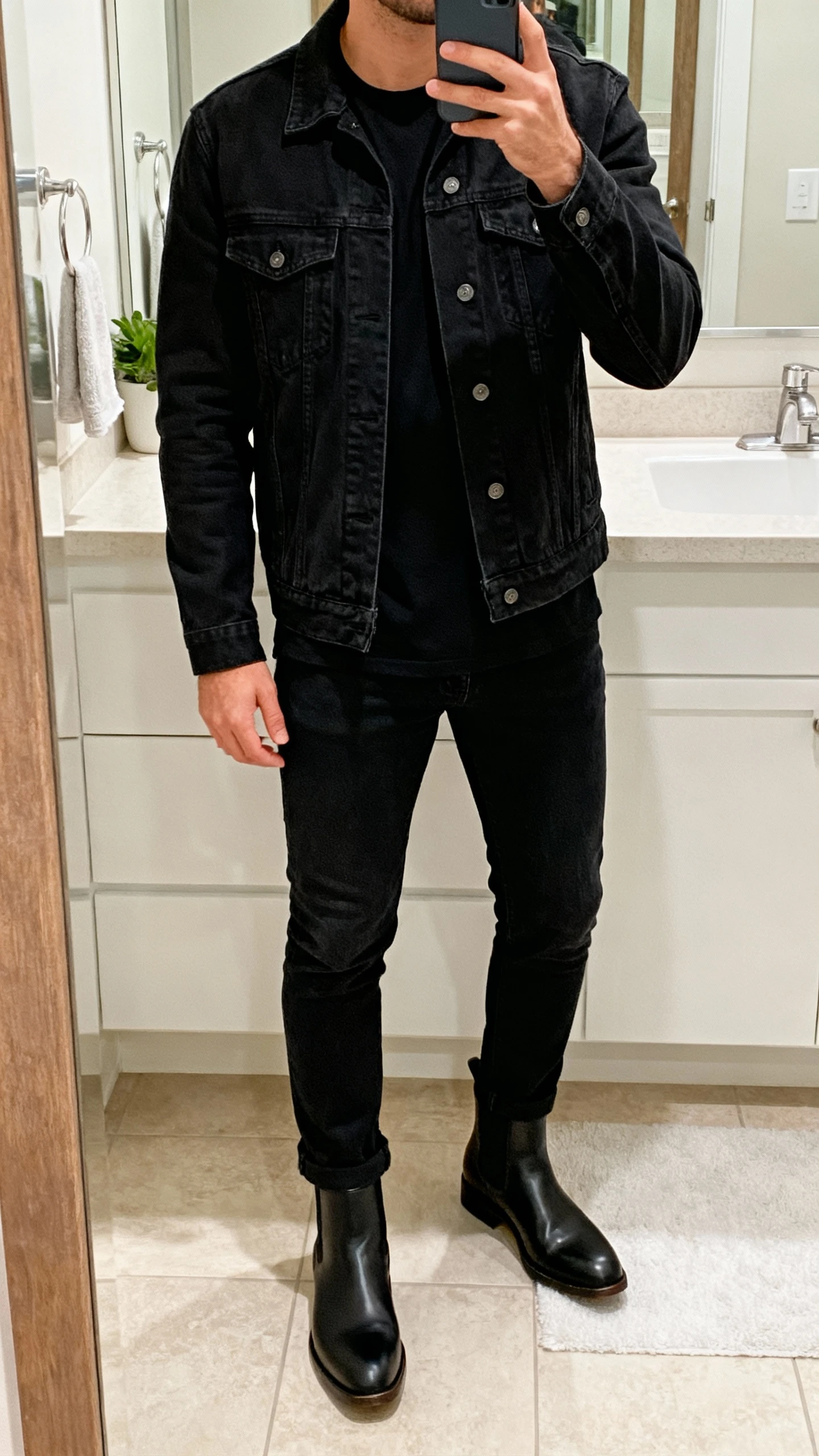 iPhone mirror selfie of a man wearing all-black: black denim jacket, black tee, black jeans, and black Chelsea boots, face not visible, bathroom mirror with soft indoor lighting, natural iPhone photo.