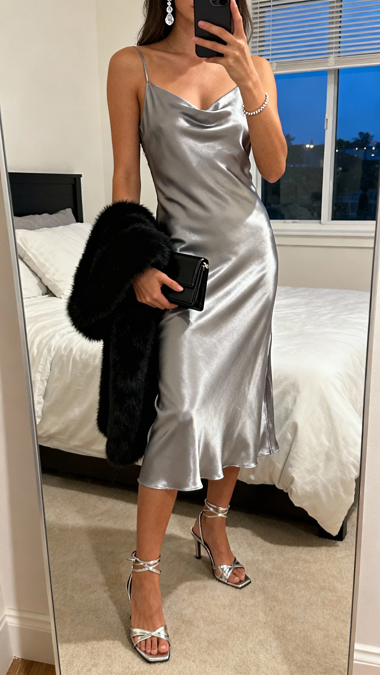 iPhone mirror selfie of a woman in a bias-cut metallic silver slip dress (midi), chrome strappy heels, structured black mini clutch, delicate crystal drop earrings, thin tennis bracelet, and a soft black faux-fur stole draped over one arm; face not visible, bedroom mirror, evening window light, natural iPhone photo quality.